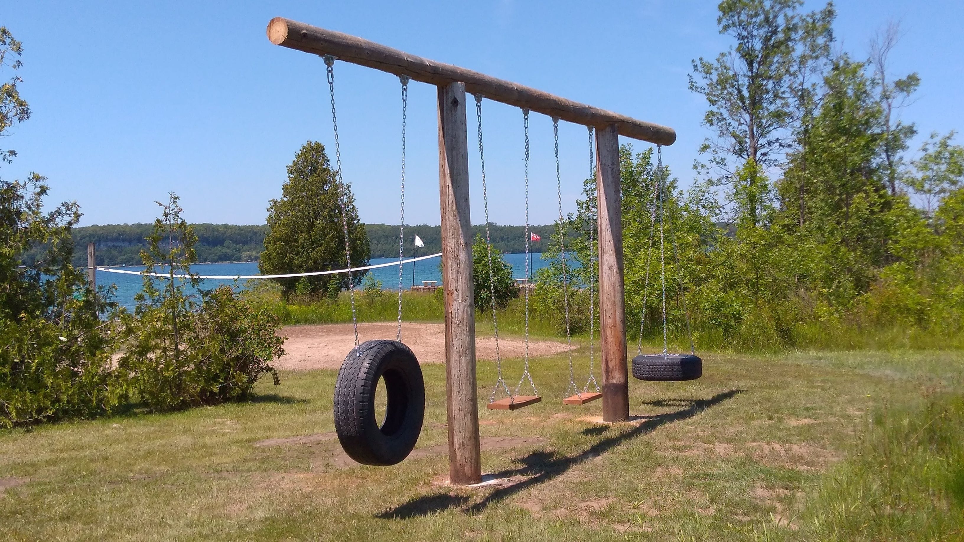 A wooden swing set with tire swings, a rope swing, and a view of a lake with trees and a bridge in the background.
