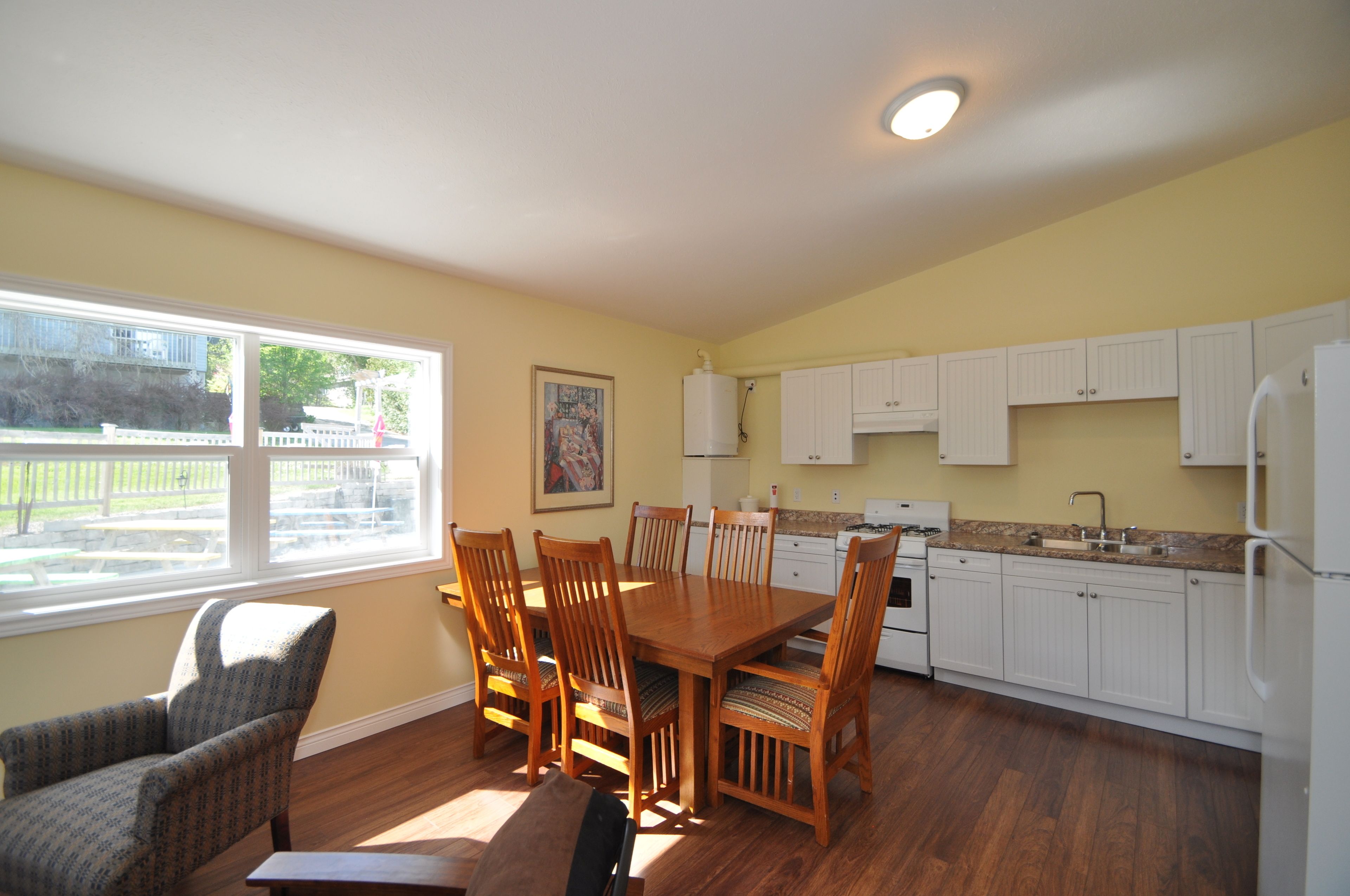 A kitchen with wooden cabinets, a dining table with chairs, a refrigerator, and a window with a view of a fence and trees.