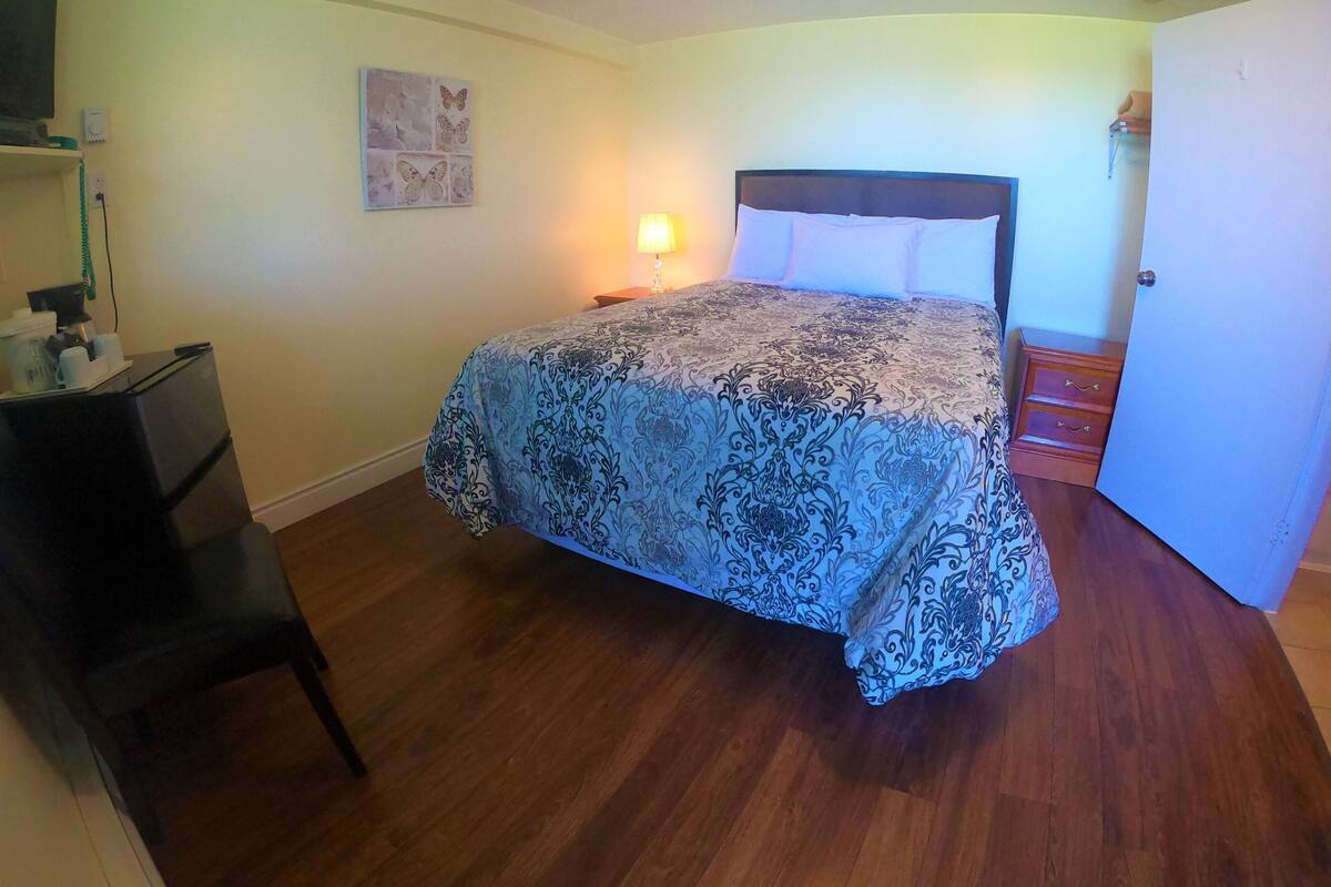 A bedroom with a bed, a desk, a chair, a nightstand, and a wall-mounted lamp.
