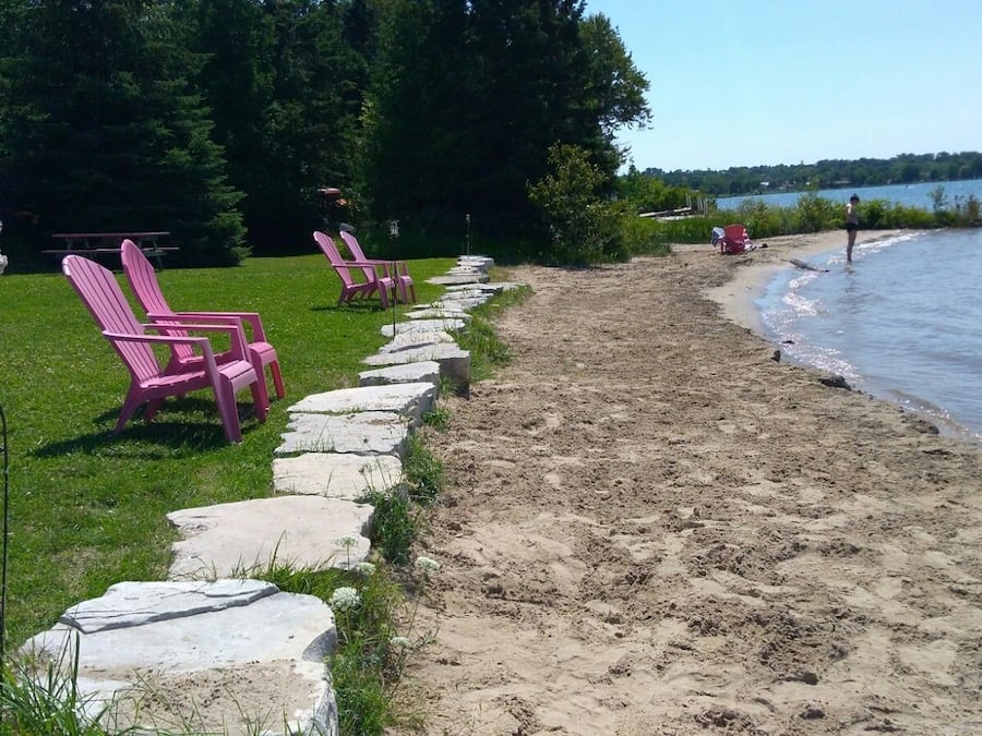 A lakeside area with a stone pathway leading to pink chairs, a picnic table, and a lamp post.