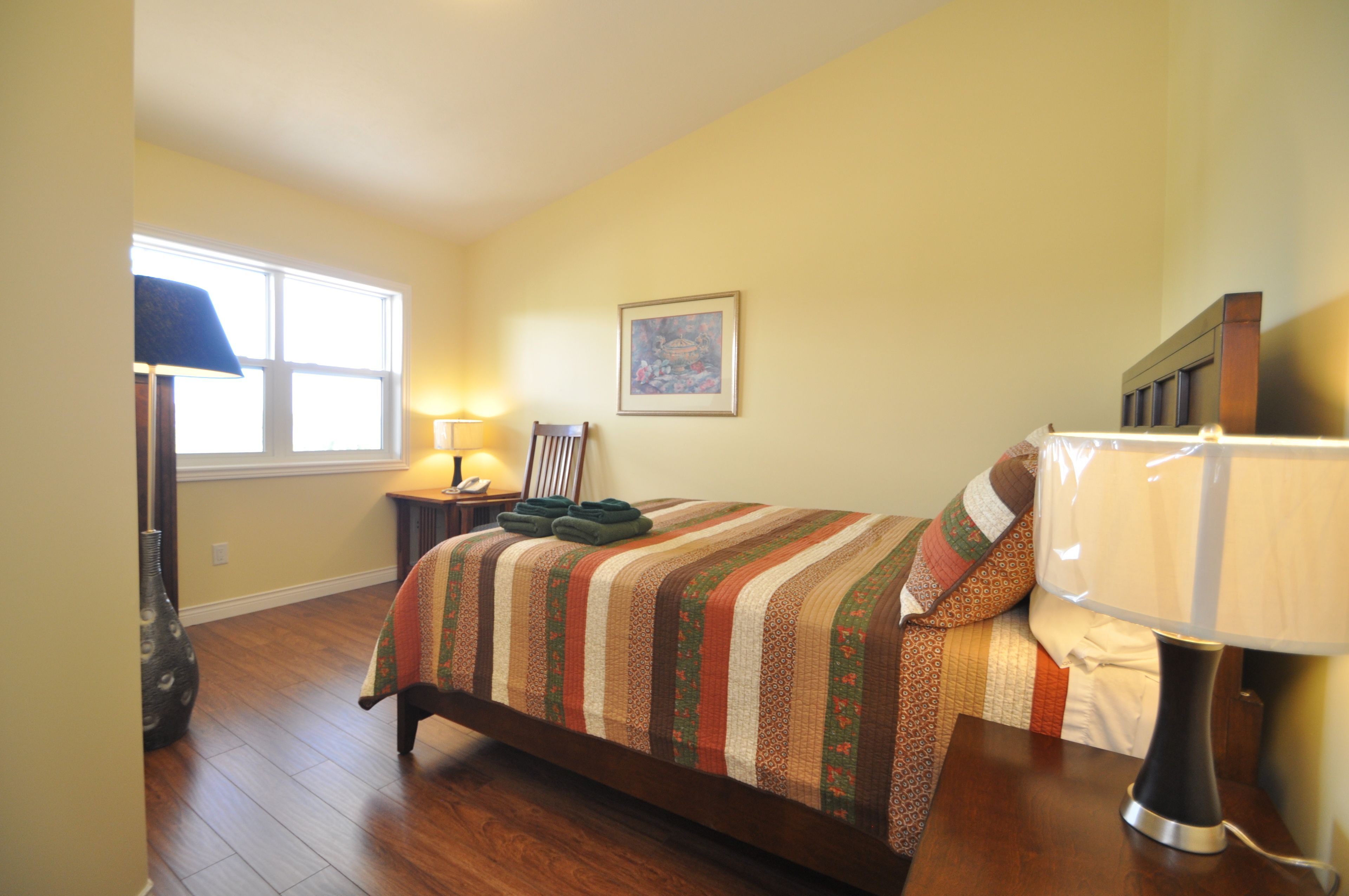 A bedroom with a bed, a nightstand, a lamp, a chair, a window, and a painting on the wall.