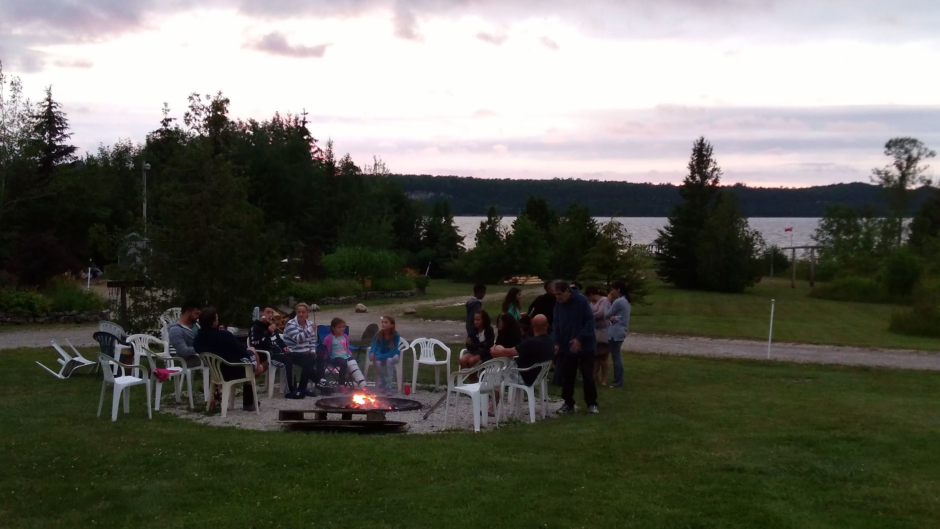 A group of people gathered around a campfire in an outdoor setting with chairs and a grassy area.