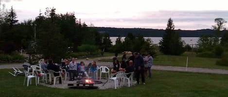 A group of people gathered around a campfire in an outdoor setting with chairs and a grassy area.