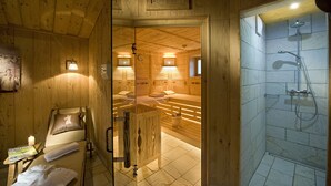 Una habitación tipo cabaña de madera con una cama de madera, un banco, una pequeña mesa con una vela y una zona de ducha con puerta de vidrio.
