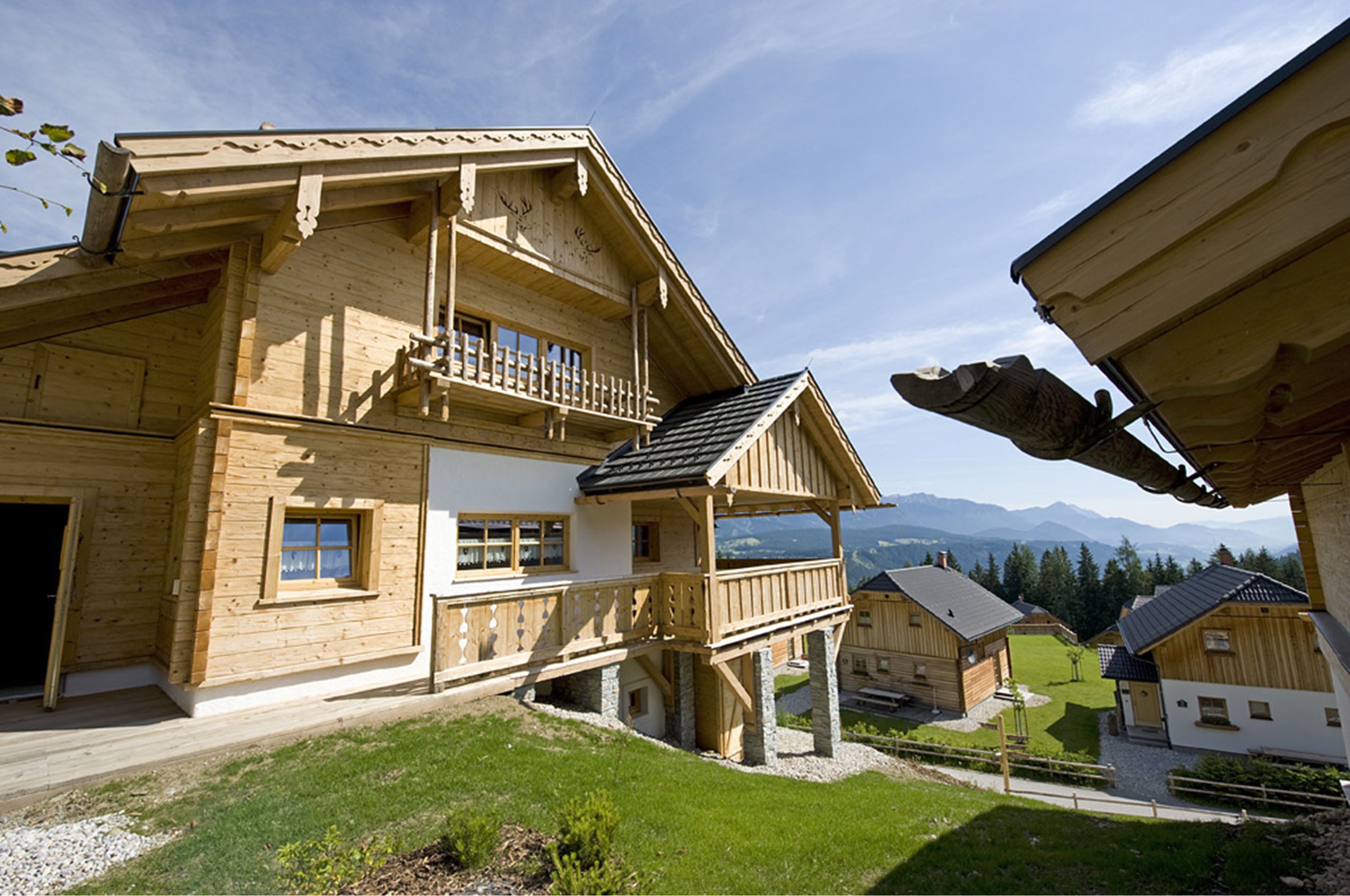 A wooden chalet with a balcony, a stone foundation, and a view of mountains in the background.