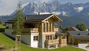 A chalet-style house with a large balcony, stone facade, and a view of snow-capped mountains. - Almdorf Reiteralm (Schladming)