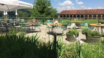 Un espace extérieur aménagé en terrasse, avec des tables et des chaises, entouré de verdure et d’un grand arbre.