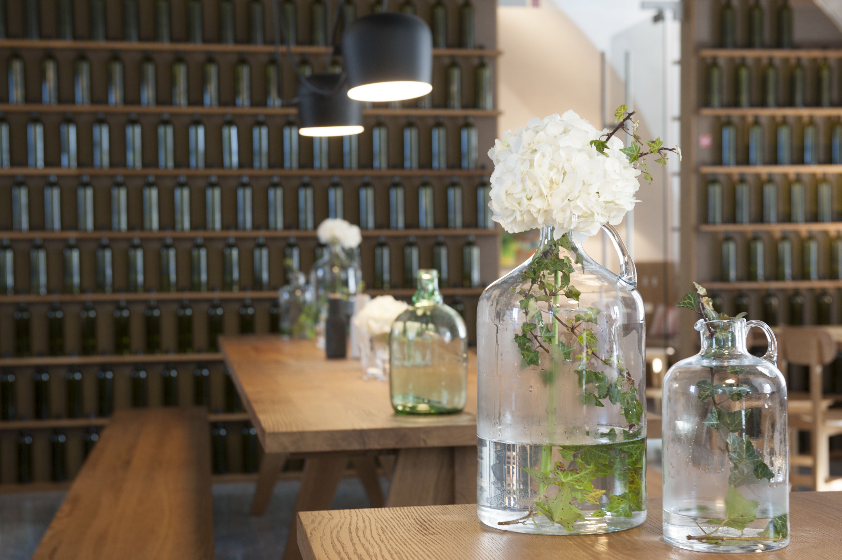 Une table en bois, sur laquelle sont disposées des bouteilles en verre transparent remplies d’eau et agrémentées de fleurs blanches, le tout placé devant une étagère remplie de bouteilles.