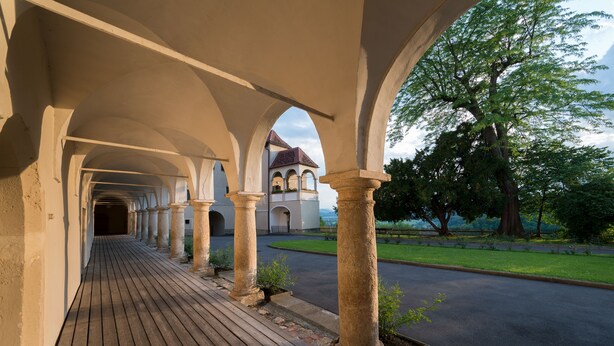 A covered walkway with stone columns, leading to a building with a red roof. - Schloss Seggau (Leibnitz)