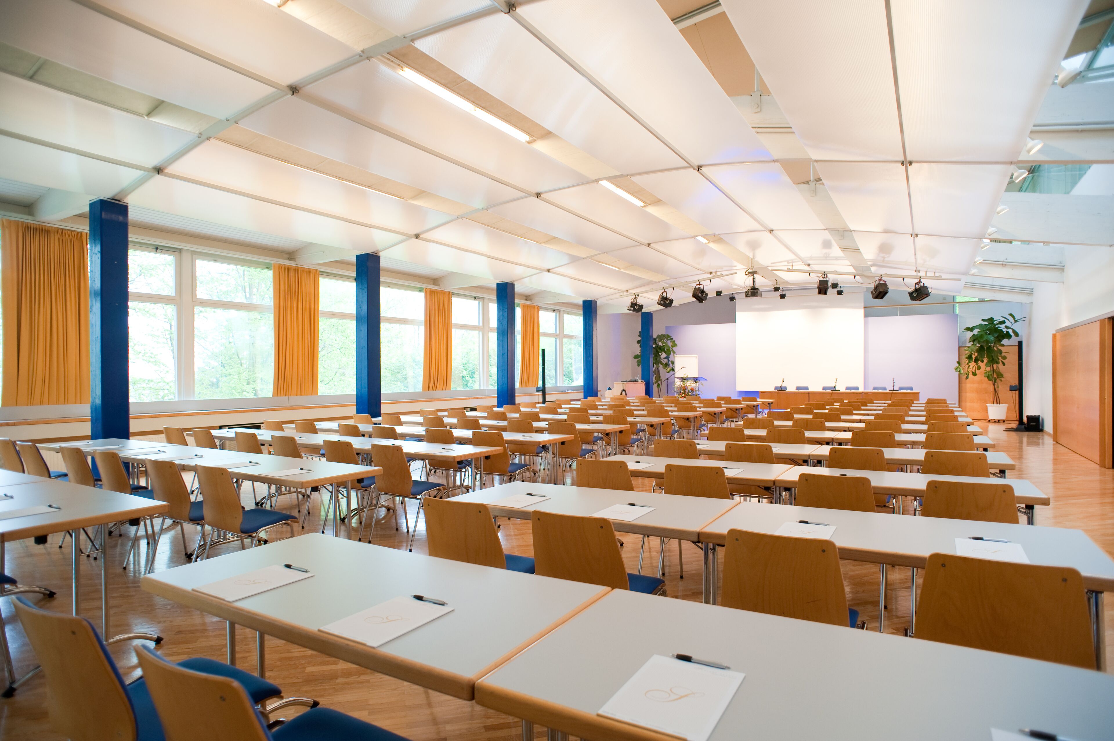 Ein Klassenzimmer mit Reihen von Tischen und Stühlen, großen Fenstern mit Vorhängen und einer Bühne mit Beleuchtung.