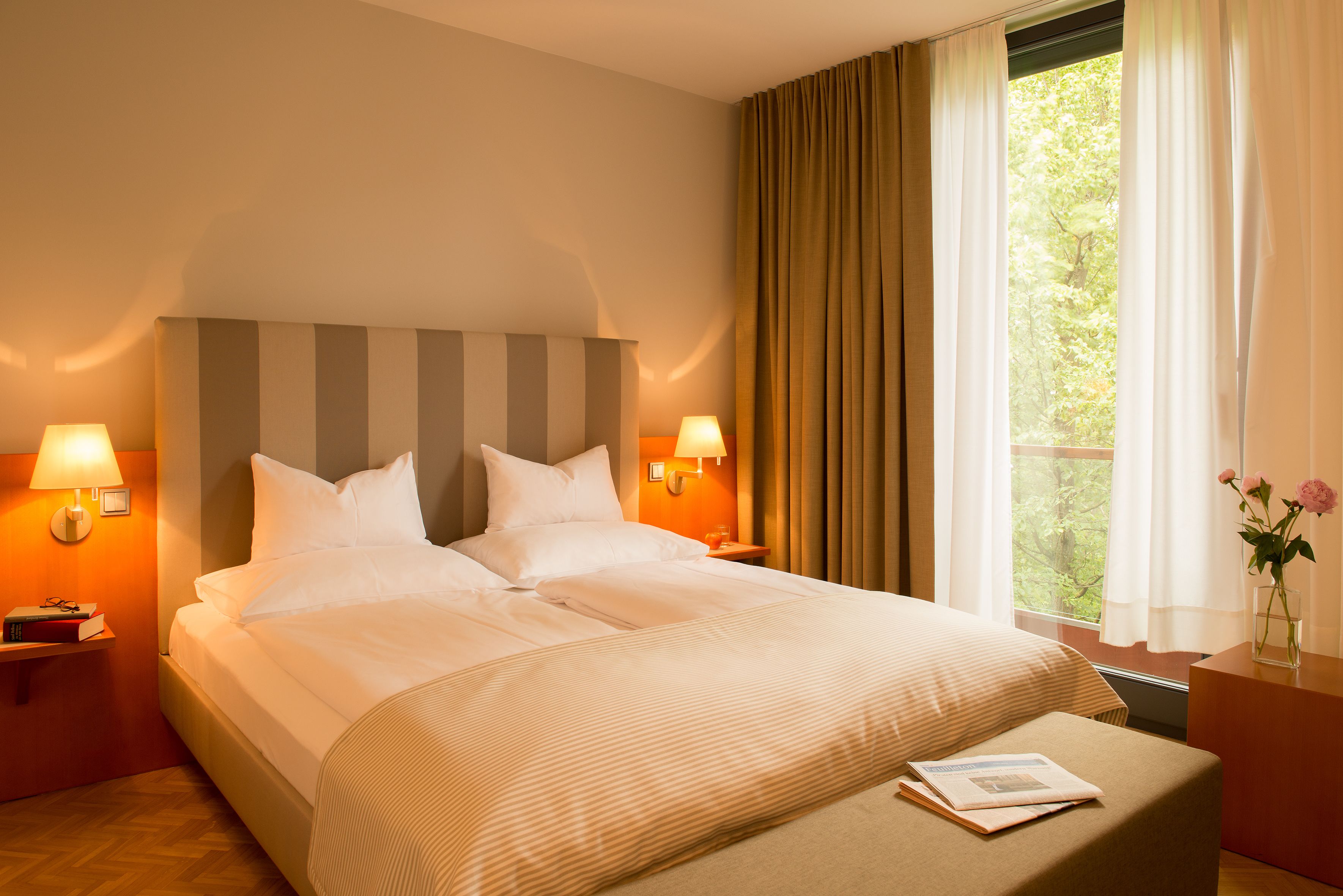 A hotel room with a large bed, two bedside lamps, a window with curtains, and a small table with a vase of flowers.
