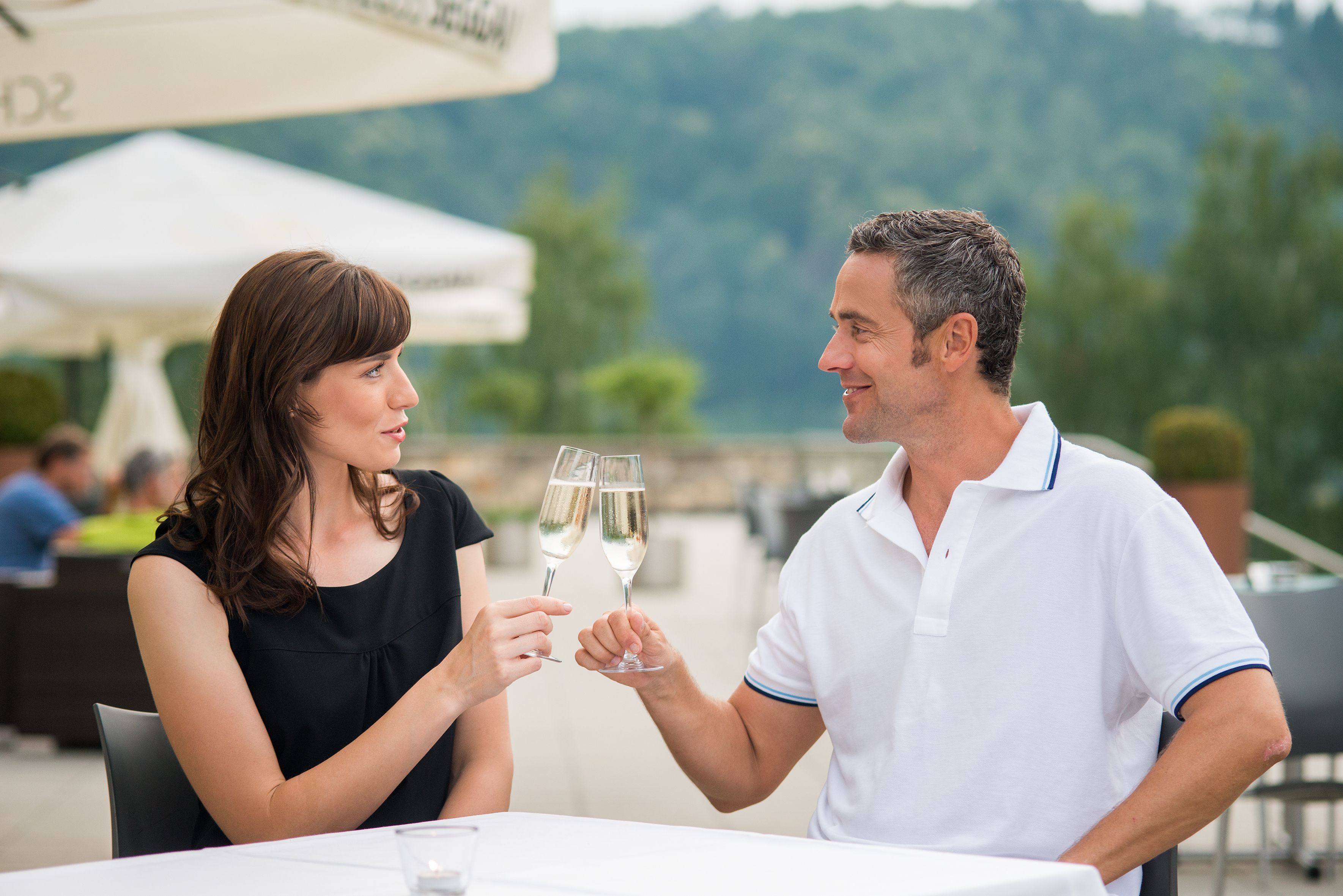 Twee personen die met champagneglazen proosten aan een buitentafel, met een witte parasol op de achtergrond.