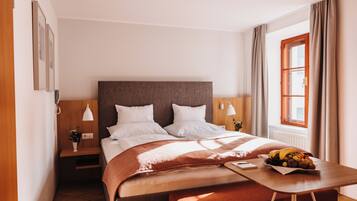 A hotel room with a bed, bedside tables, a nightstand, a window with curtains, and a fruit basket on the nightstand.