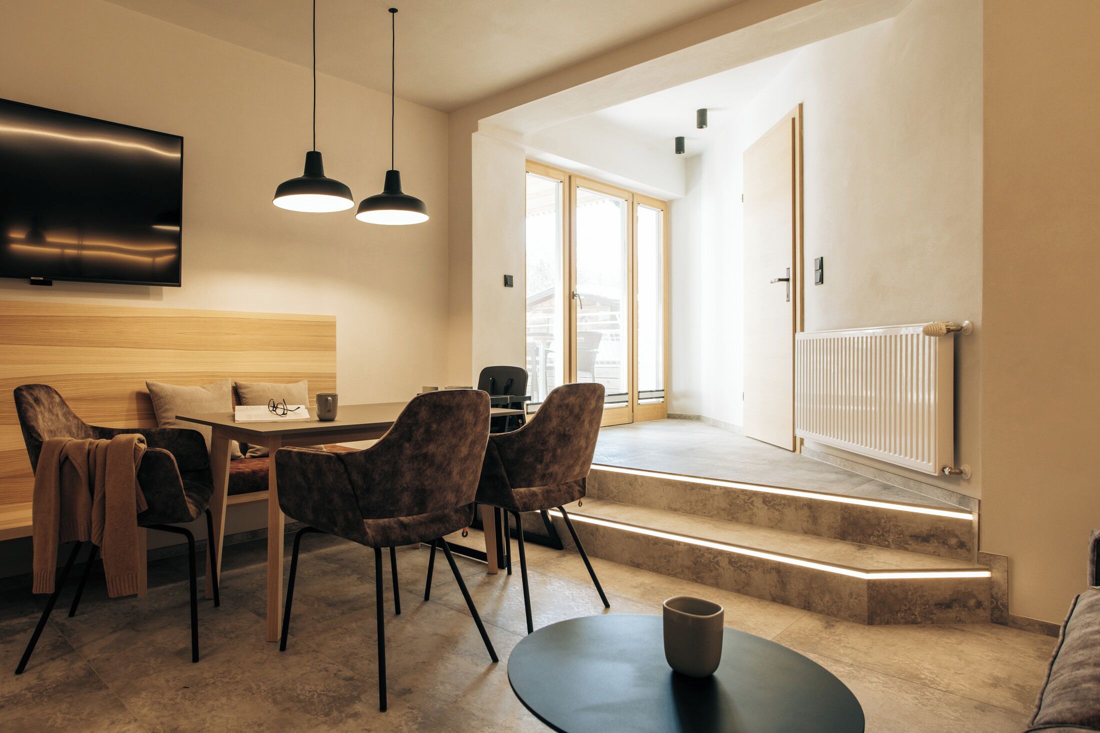 A modern dining area with a wooden table and chairs, a television mounted on the wall, and a staircase leading to an outdoor area.