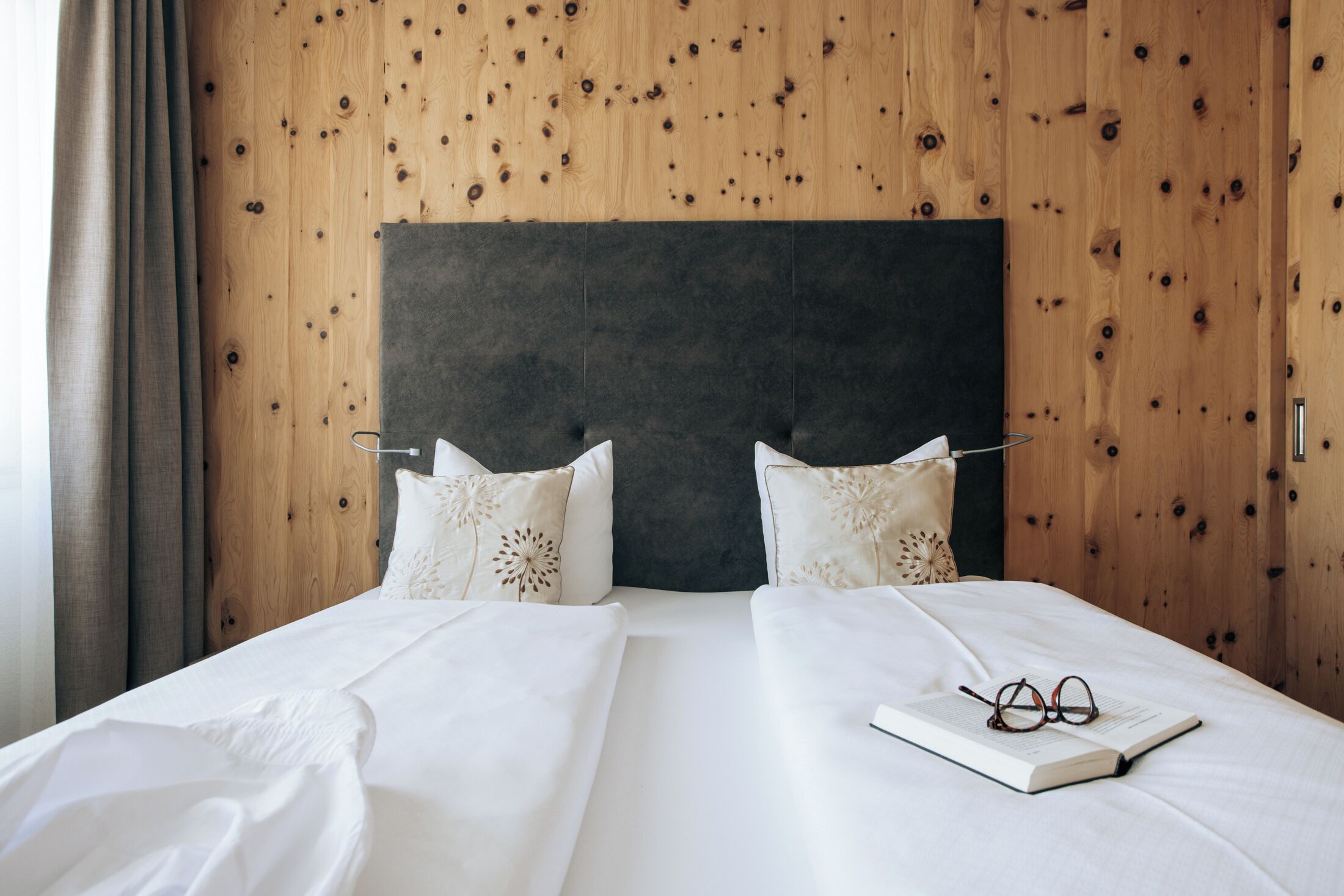 Una cama doble con un cabecero oscuro, dos almohadas decorativas y un par de gafas sobre un libro en la cama.