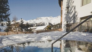 Una piscina cristalina con muelle de madera, rodeada de montañas nevadas y un edificio de fachada blanca.