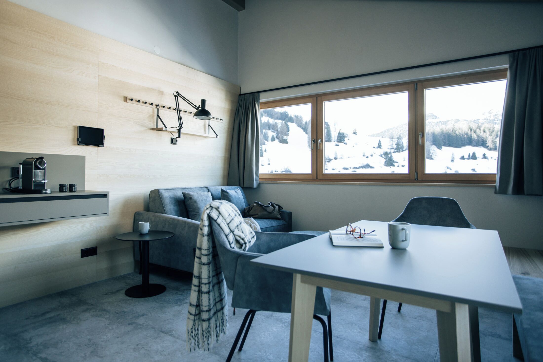 A modern living room with a sofa, a dining table, and a view of a snowy landscape through large windows.