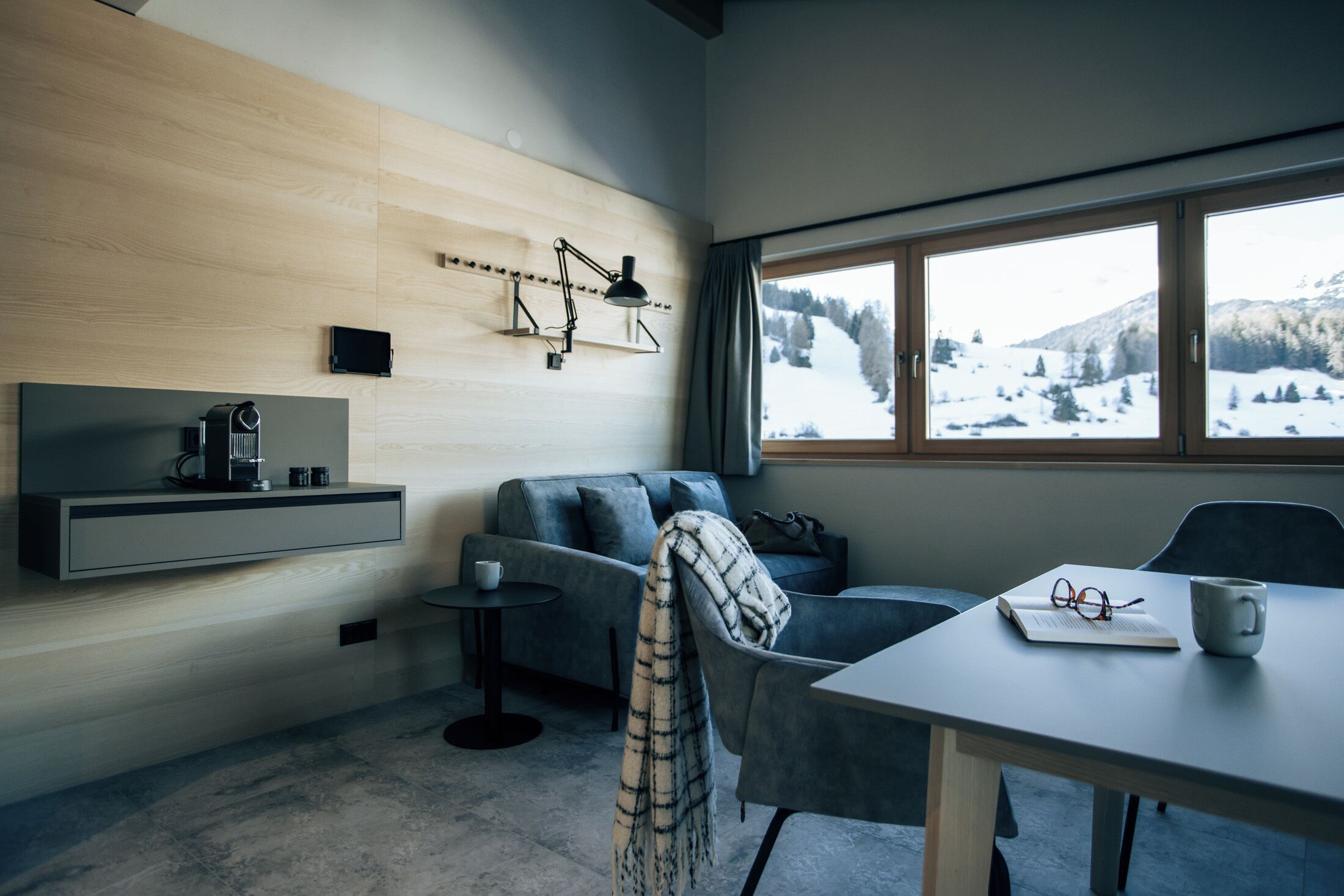 Ein modernes Wohnzimmer mit einem grauen Sofa, einem kleinen Tisch mit Tasse und Gläsern, einer Wandlampe und einem Fenster mit Blick auf Schnee.