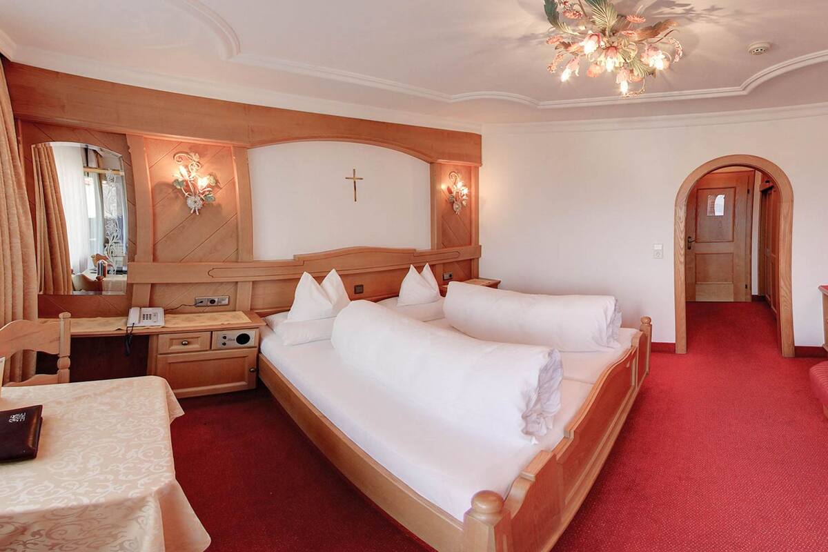 Une chambre d’hôtel avec un grand lit, un bureau, une chaise et une petite table. On y trouve un miroir, un crucifix au mur et un lustre.