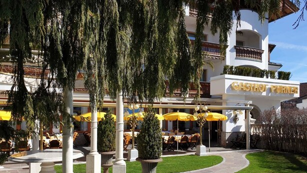 Ein Hotel mit Außenbestuhlung unter gelben Sonnenschirmen, einem gepflegten Rasen und einem Gebäude mit der Aufschrift "GASTROF PUNER".
