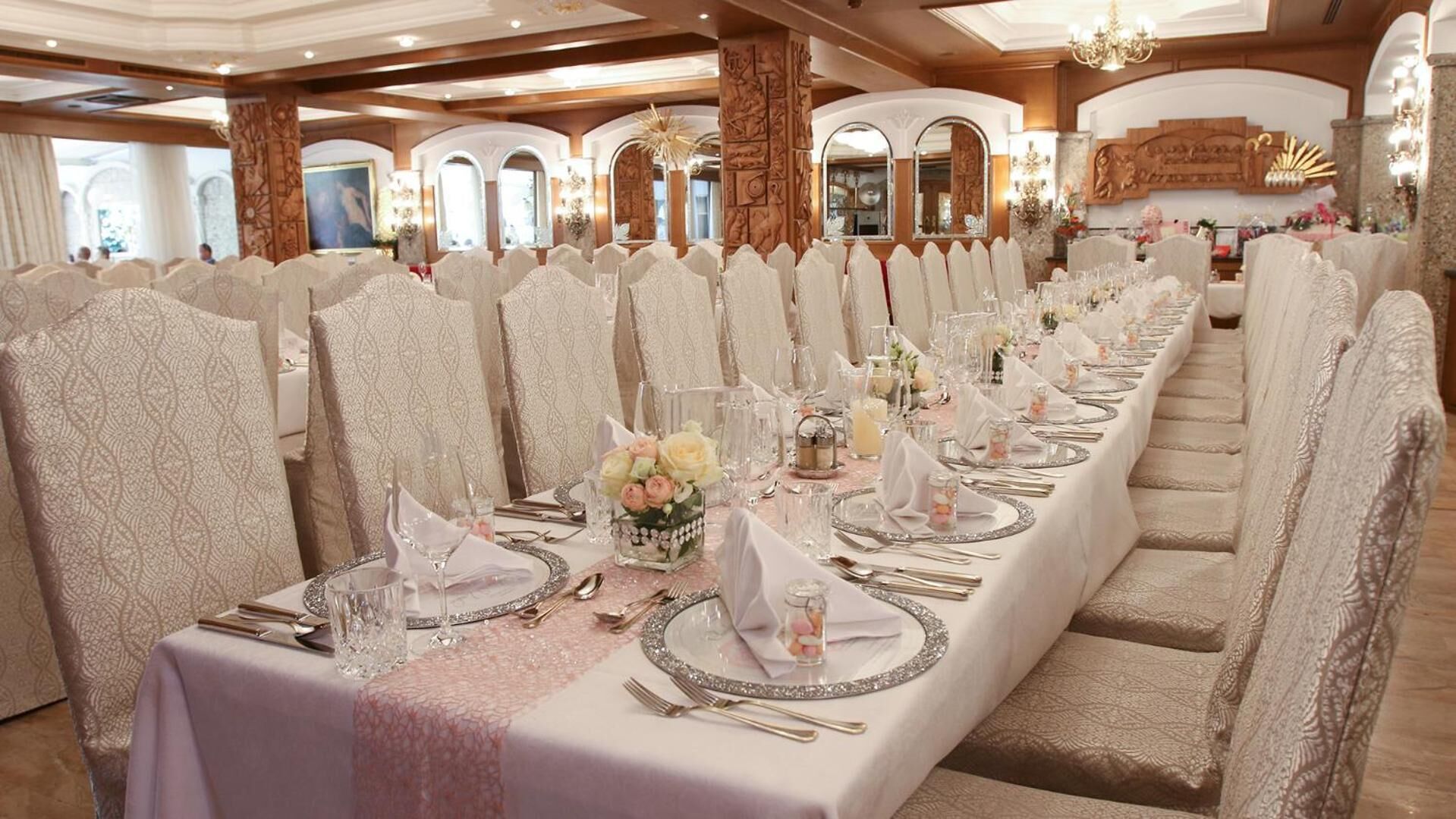 Uma longa mesa de banquete posta para um evento formal, com toalhas brancas, taças de cristal e centros de mesa florais.