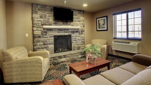 A living room with a stone fireplace, a flat-screen TV mounted above it, and a large window with blinds. - Cobblestone Inn & Suites - Harper (Harper)
