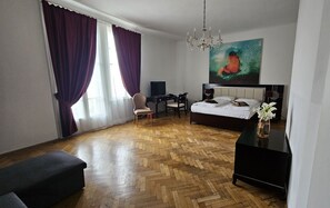 Habitación de hotel con piso de madera en espiga, una cama, un escritorio con silla, un televisor y un cuadro en la pared.