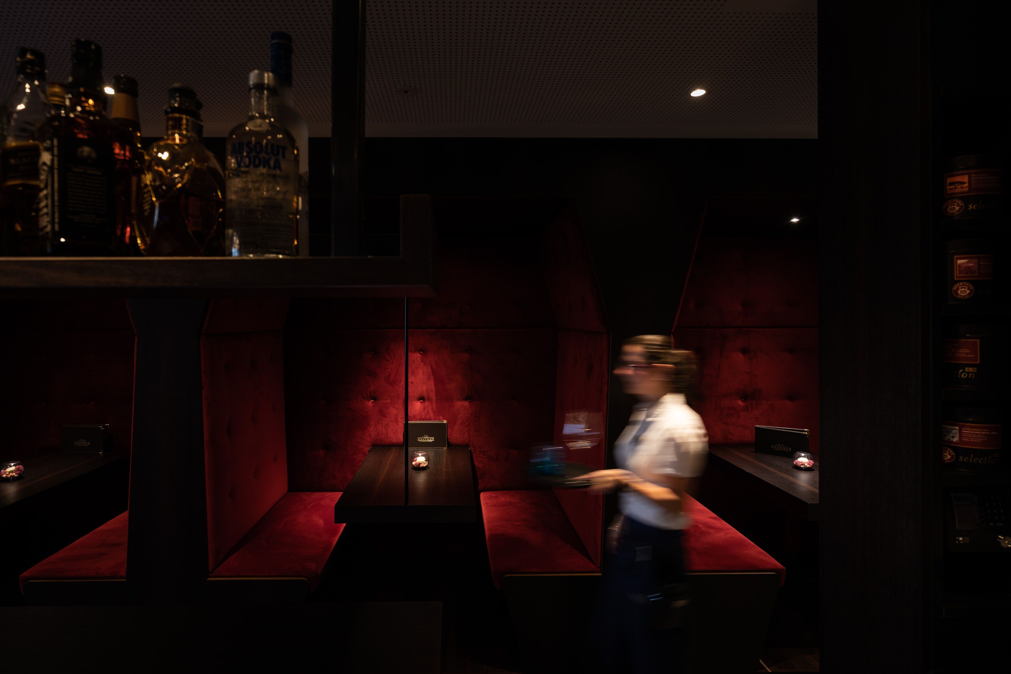 Zona bar scarsamente illuminata, con sedute imbottite rosse e diverse bottiglie di liquore sugli scaffali.