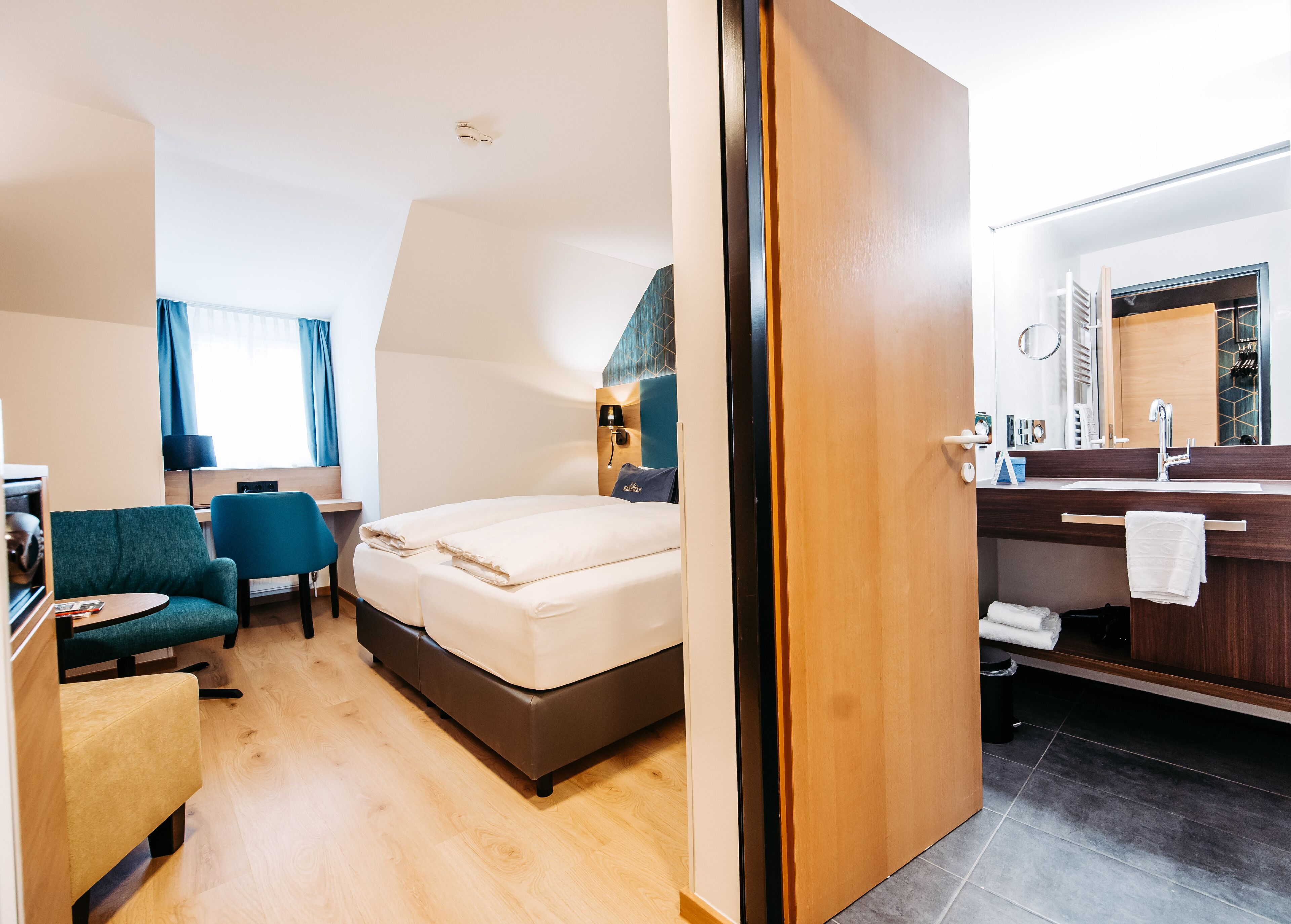 A hotel room with a bed, a desk, a chair, a small table, and a bathroom with a sink and mirror.