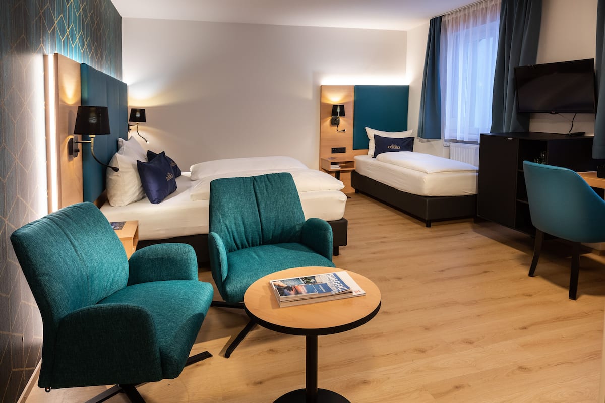 A modern hotel room with a bed, two teal armchairs, a small wooden table, a TV, and a desk.