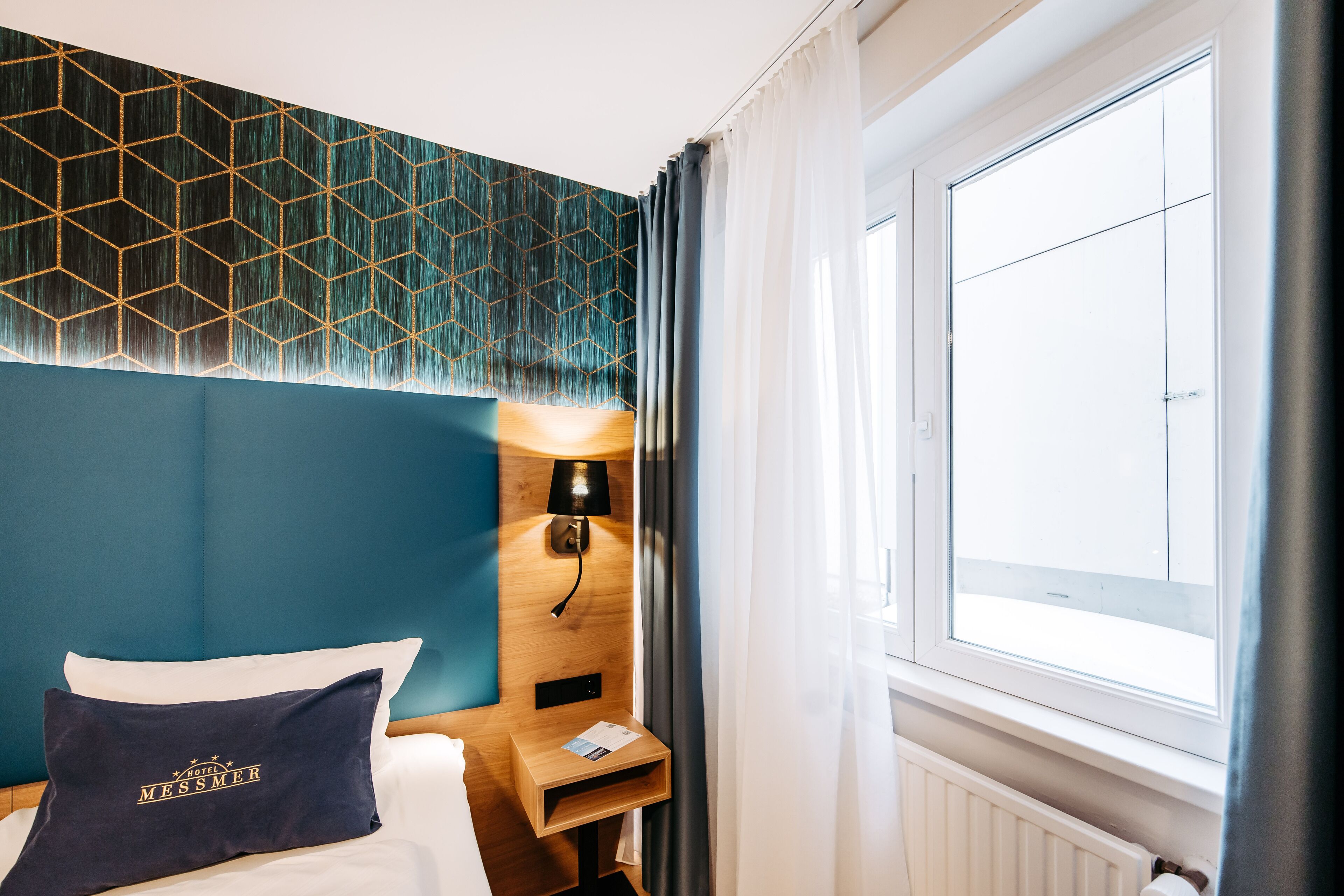 Ein Hotelzimmer mit einem Bett, einem Nachttisch, einer Lampe, einem Fenster mit Vorhängen und einer Wand mit geometrischem Muster.