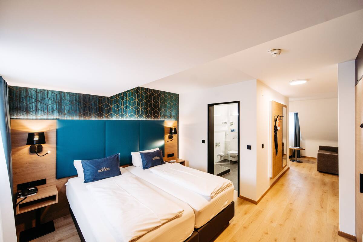 A modern hotel room with a large bed, a headboard with geometric patterns, bedside lamps, and a view into the bathroom.
