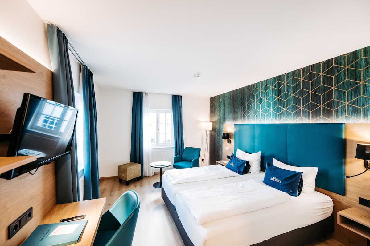 A hotel room with two beds, a desk, a TV, and a window with curtains.