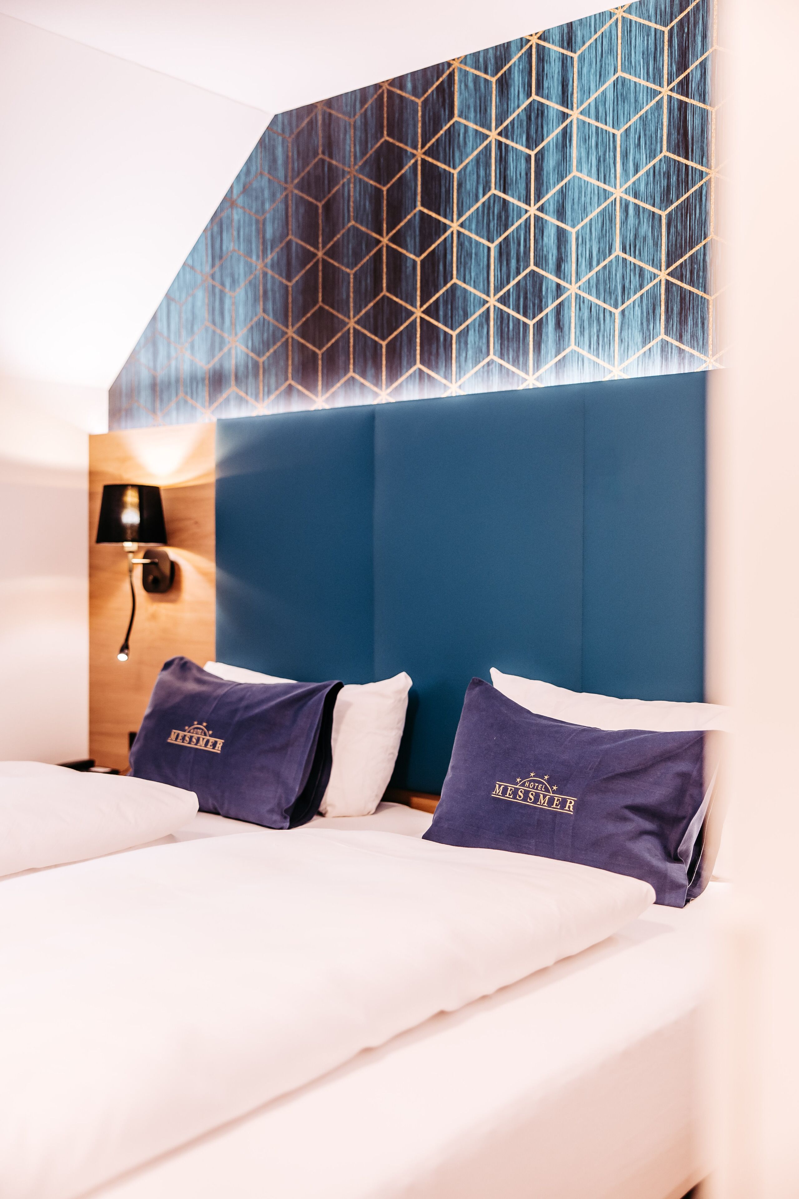 A hotel room with a modern design, featuring a large bed with white linens, two dark blue pillows, and a geometric patterned headboard.