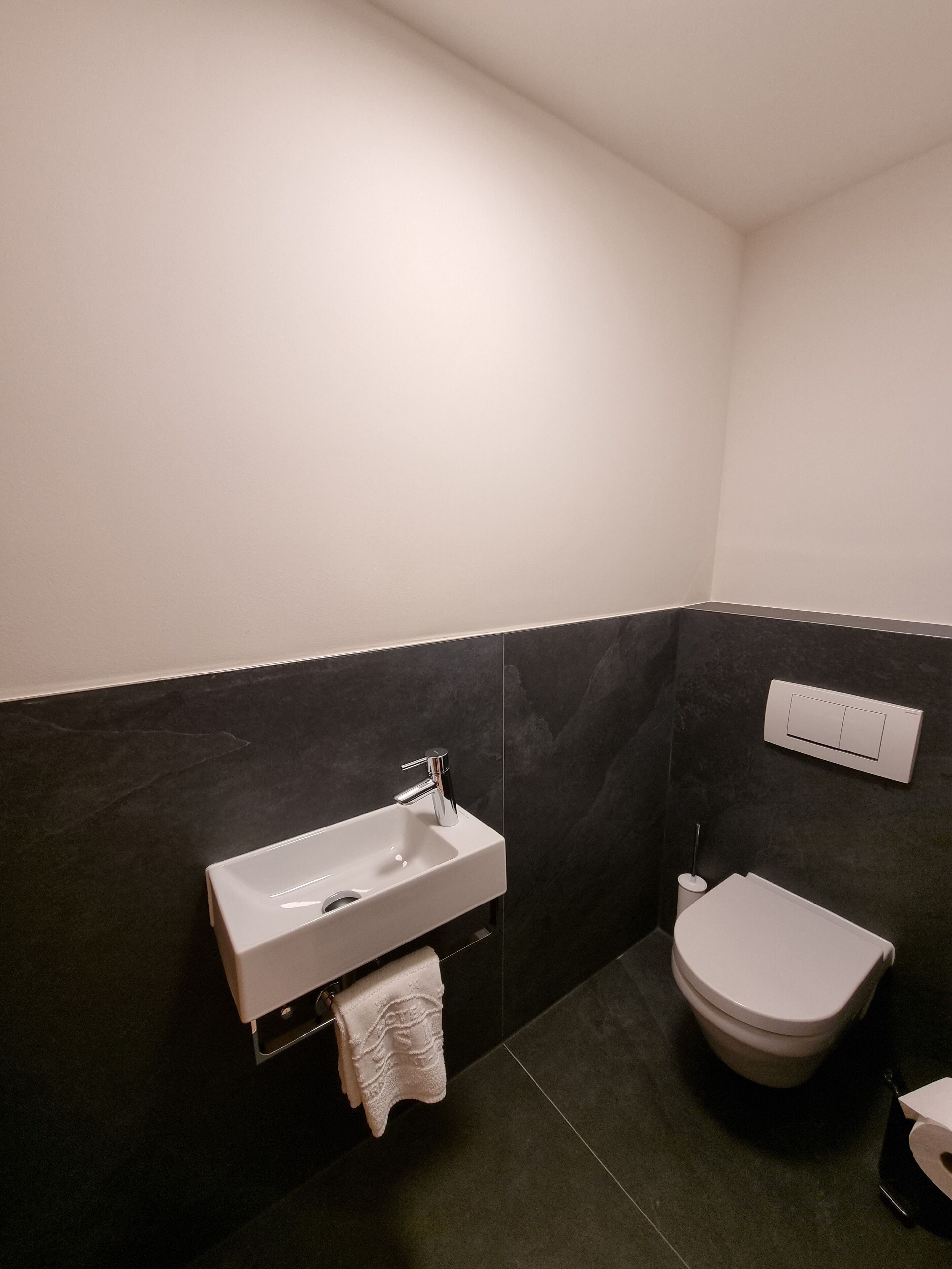Un baño moderno con un lavamanos blanco, paredes cubiertas de baldosas oscuras y un inodoro blanco.