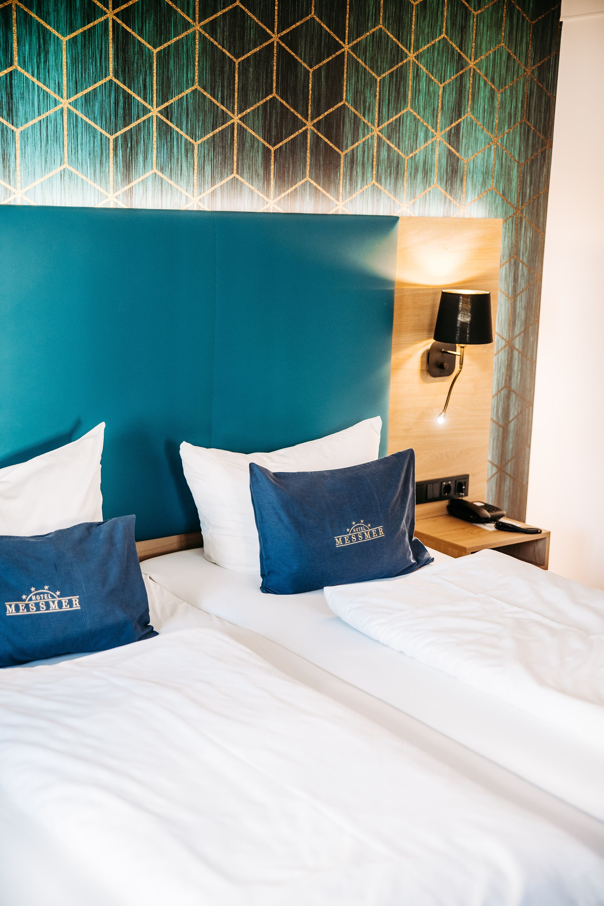 Habitación de hotel con cama, dos almohadas, mesita de noche, lámpara y pared con un diseño geométrico.