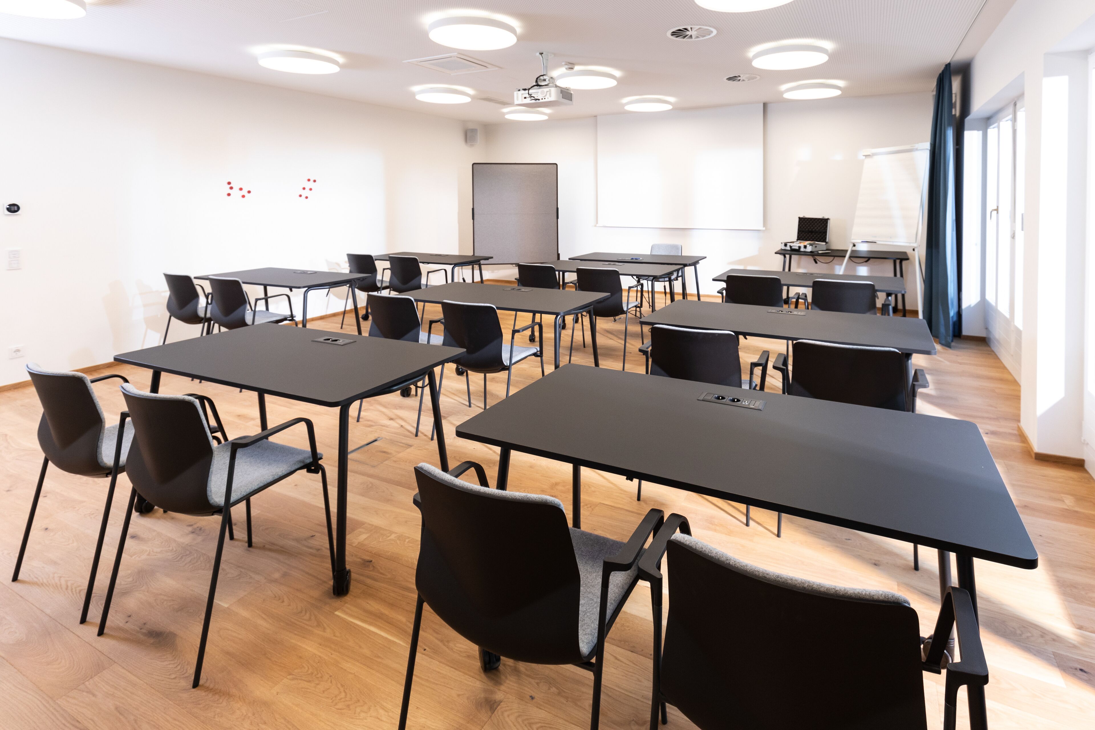 Un salón de conferencias con varias mesas y sillas, una pizarra y una pantalla de proyector.