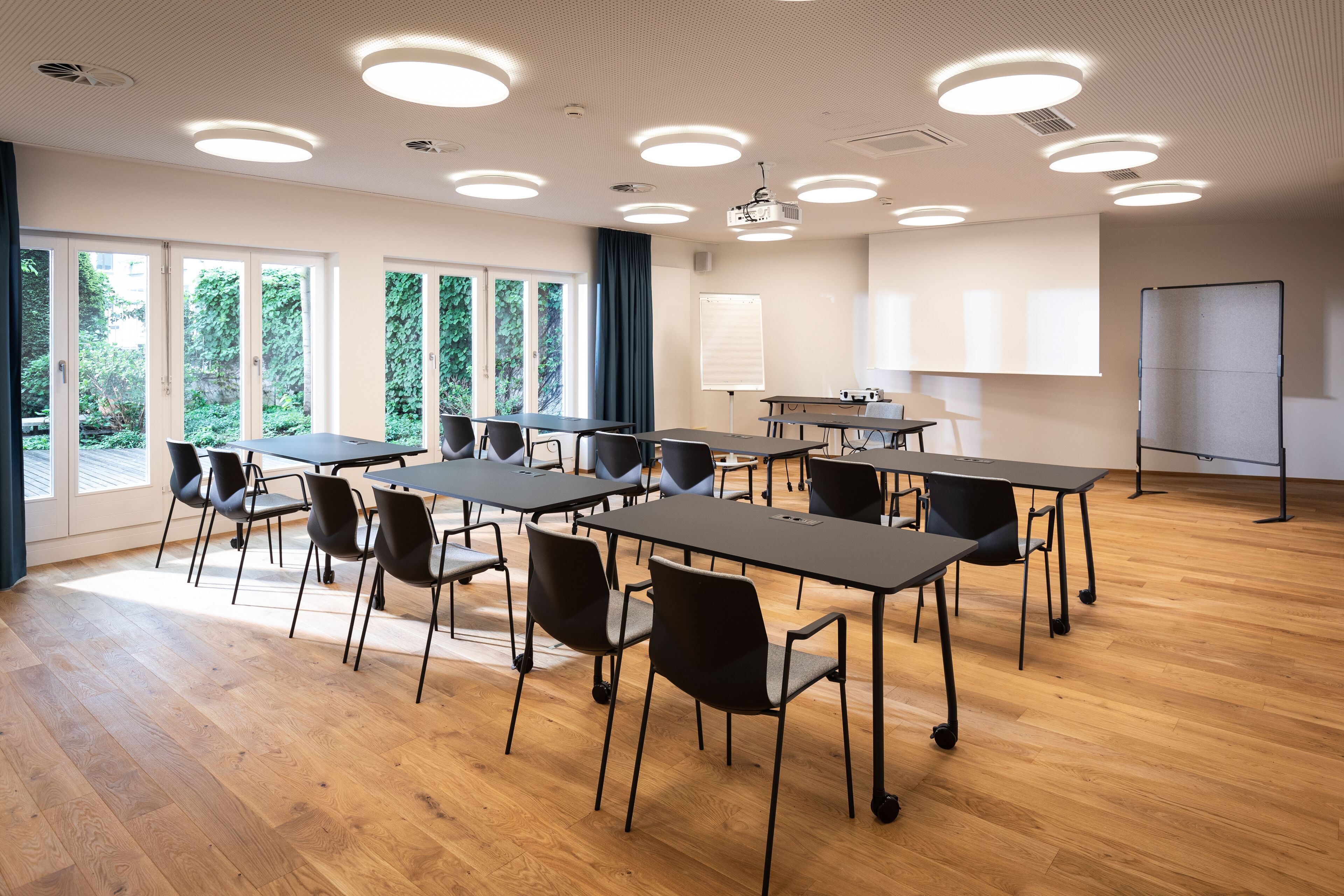 Un salón de conferencias con varias mesas y sillas, amplias ventanas que dan a zonas verdes, una pizarra y un proyector.