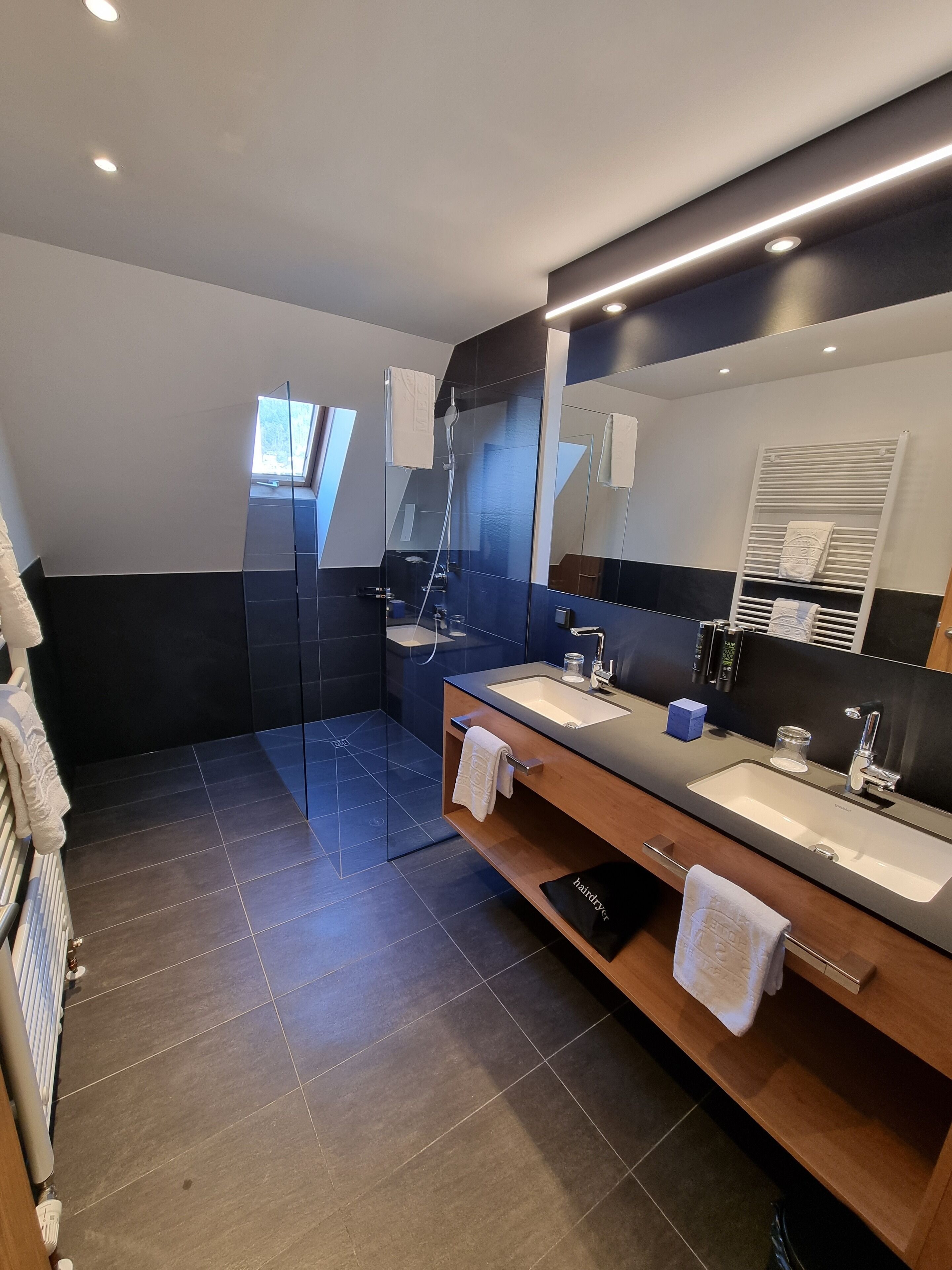 Un baño moderno con un espejo grande, dos lavamanos, una ducha y un perchero para toallas.