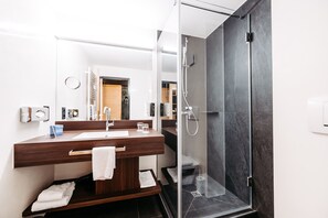 A modern bathroom with a wooden vanity, a glass shower enclosure, and a visible door to another room. - Hotel Messmer (Bregenz)
