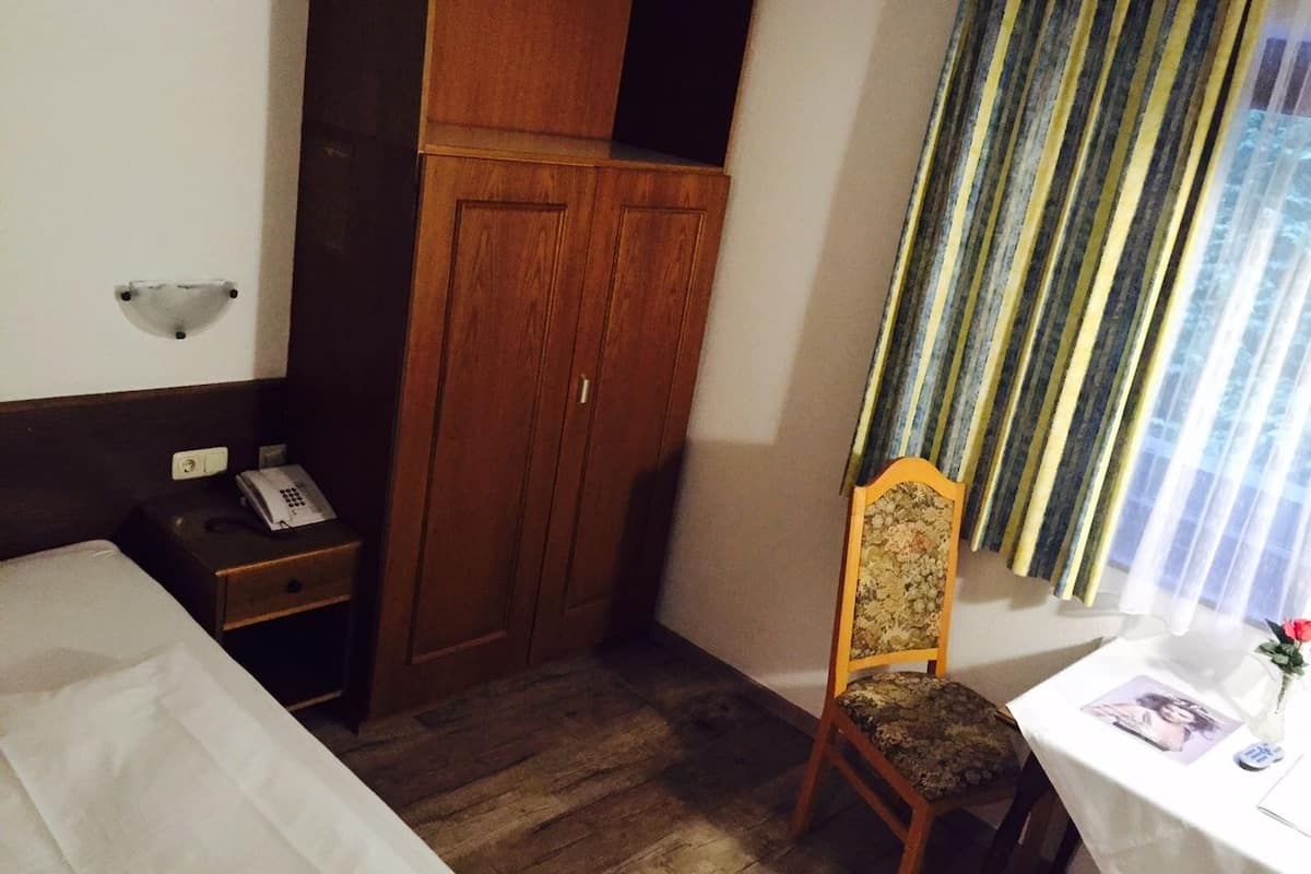A hotel room with a bed, a wooden wardrobe, a chair, a small table with a vase, and a window with curtains.