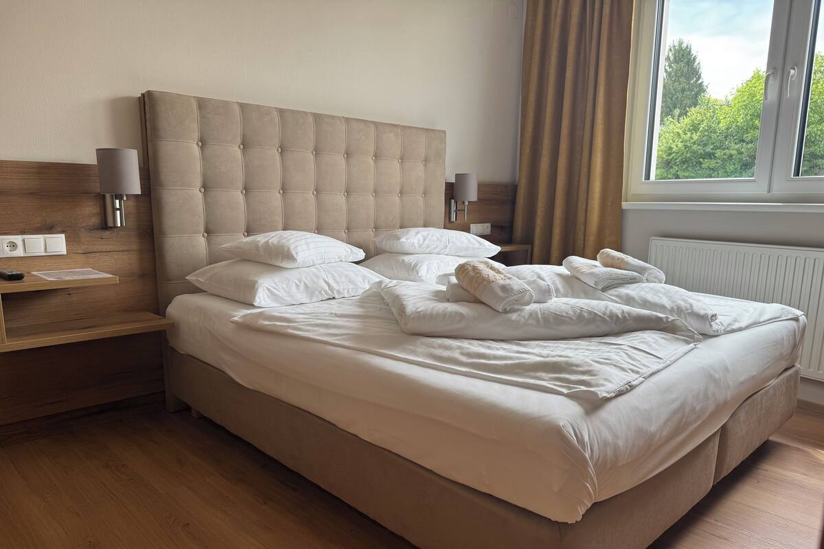 Una cama bien tendida con sábanas y almohadas blancas, un cabecero de madera, lámparas de noche y una ventana con cortinas.