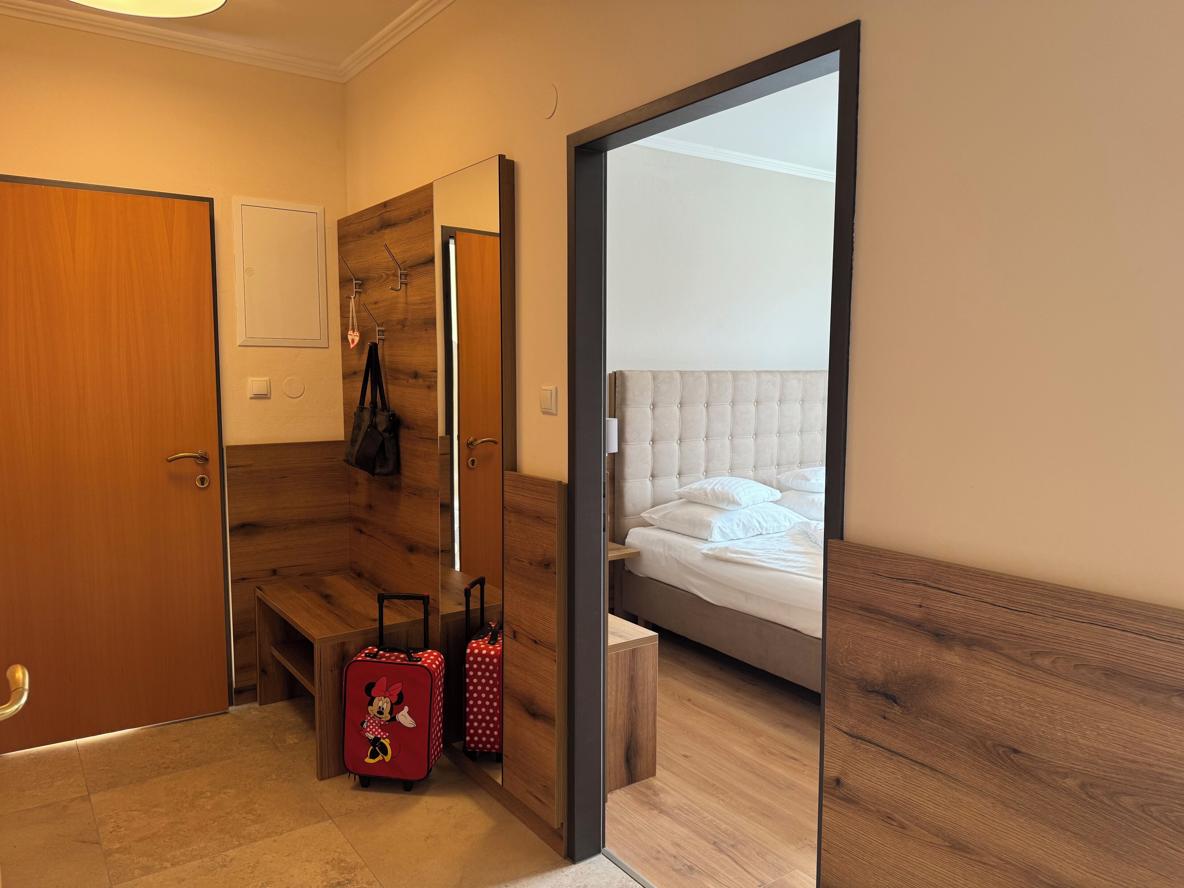 Habitación de hotel con piso de madera, una cama con ropa de cama blanca, un banco de madera y una maleta.