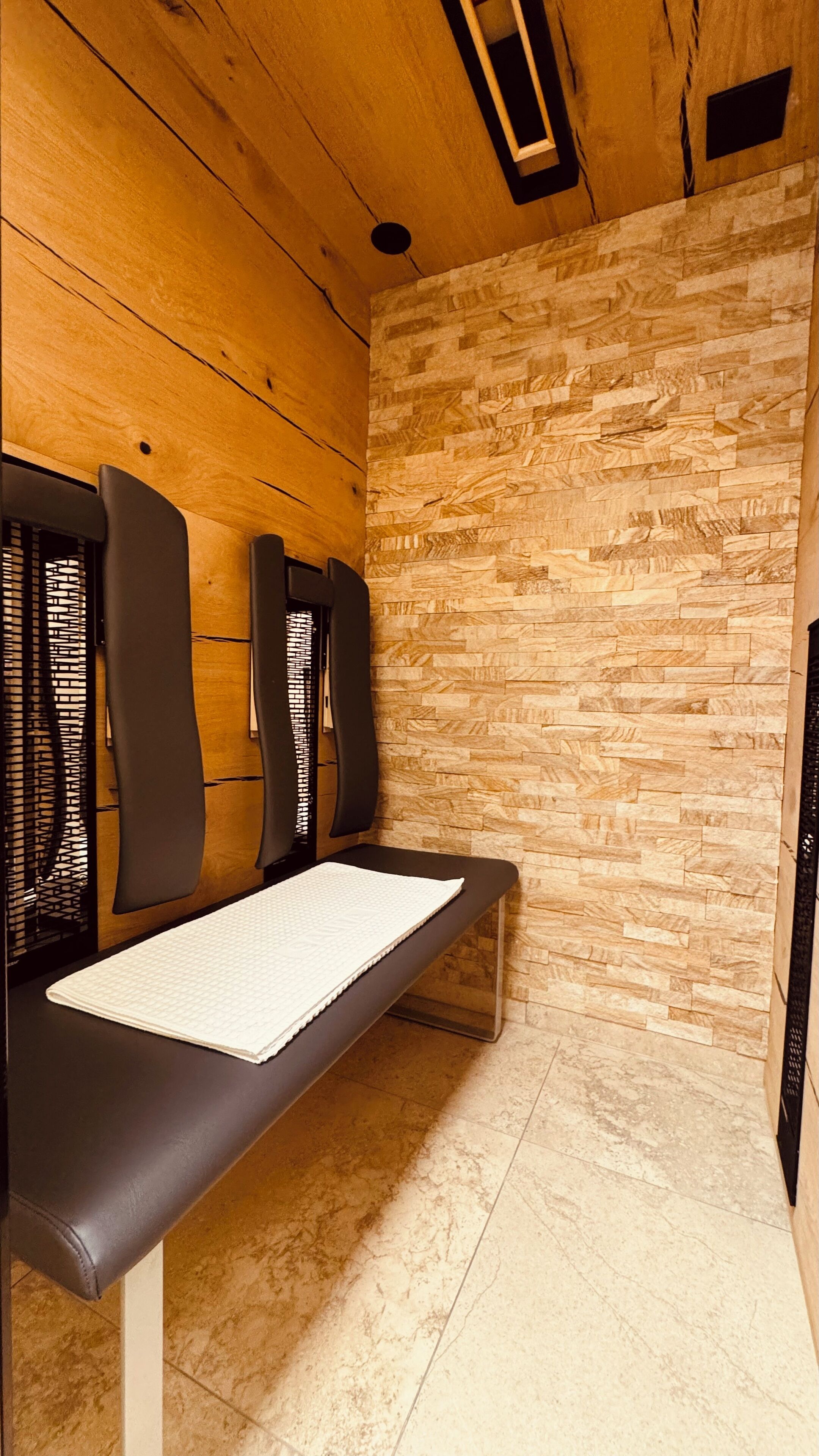 A wooden ceiling with a recessed light, a bench with a white towel, and a stone-patterned wall.
