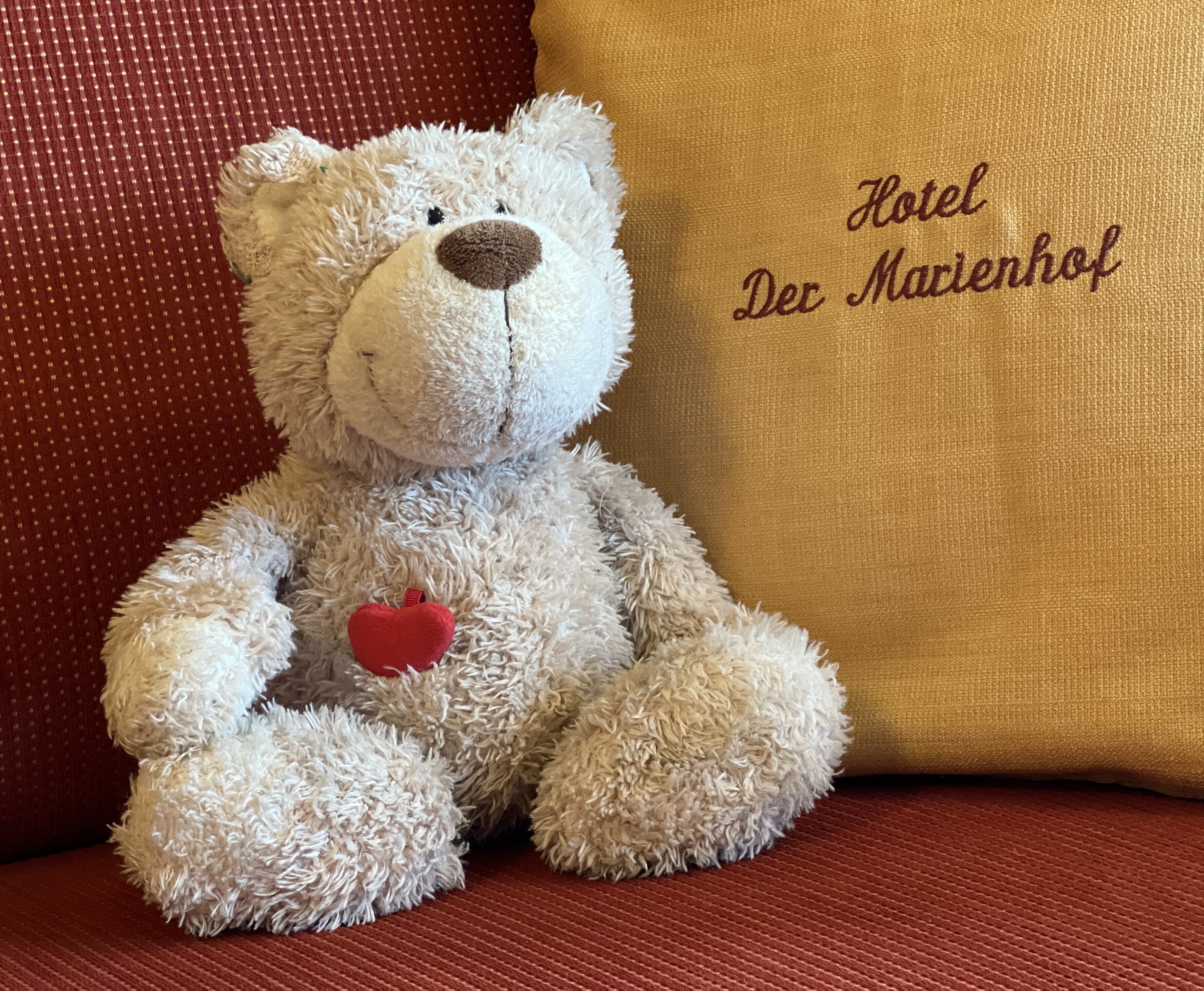 Een zachtaardig teddybeer met een hart op schoot, zittend op een rode, gestructureerde bank naast een kussen met 'Hotel Der Marienhof' geborduurd erop.
