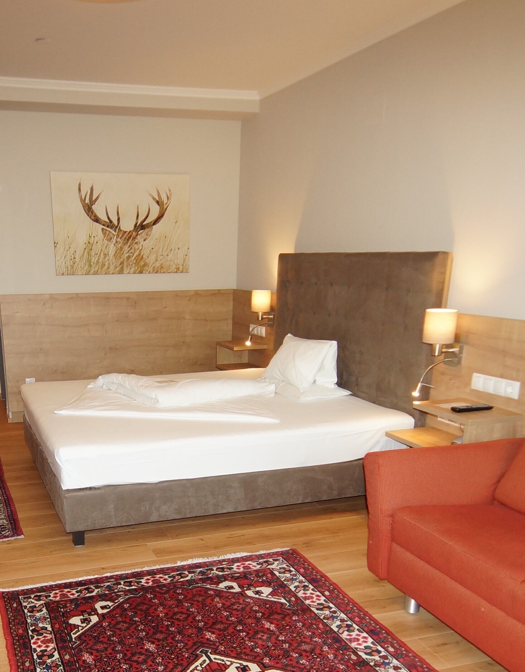 A hotel room with a large bed, two bedside tables with lamps, a red sofa, and a wall-mounted painting of deer antlers.