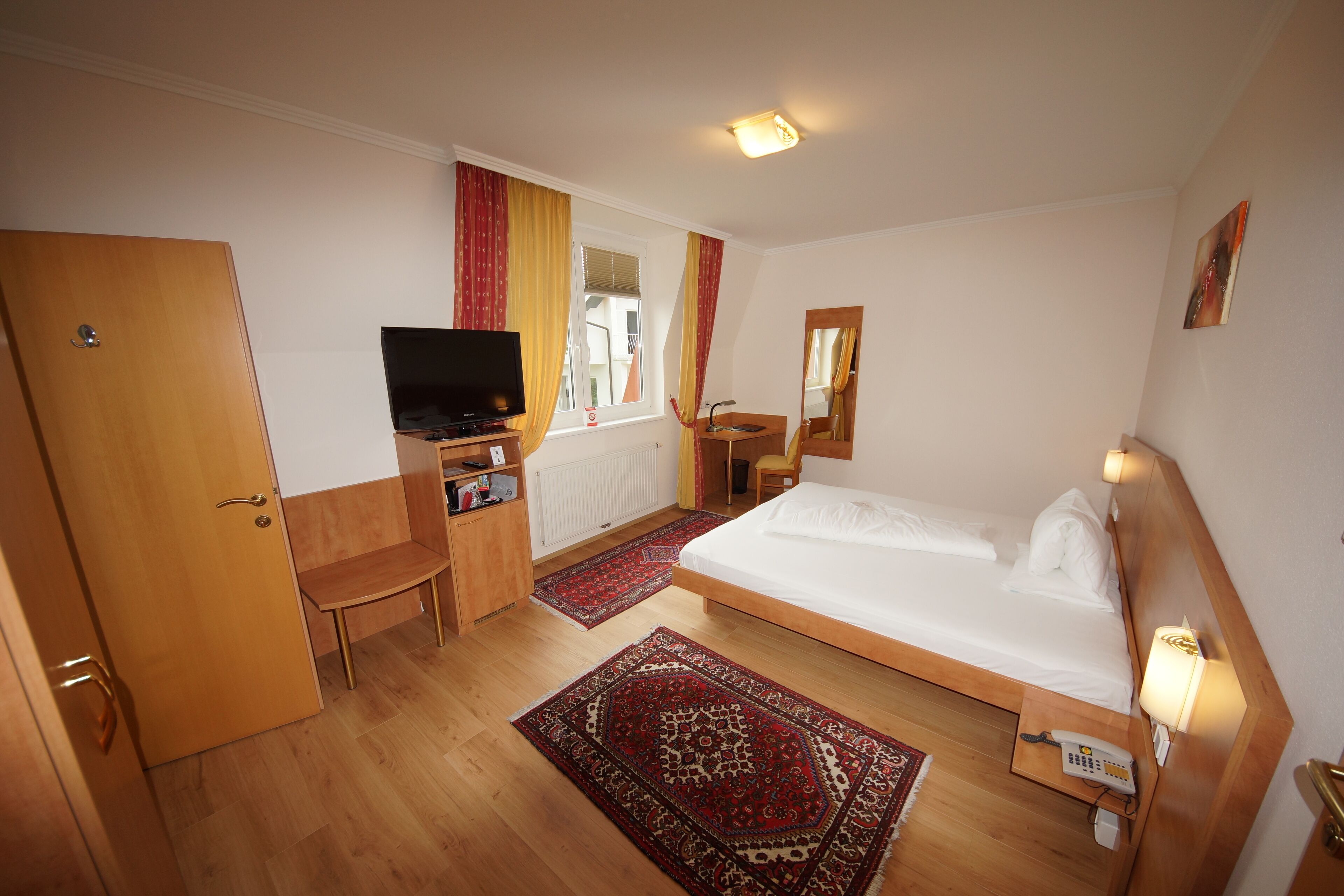 A hotel room with a bed, a desk with a computer, a TV, a chair, a small table, and a window with curtains.