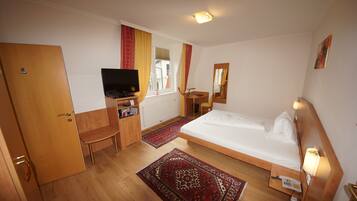 A hotel room with a bed, a desk with a computer, a TV, a chair, a small table, and a window with curtains.