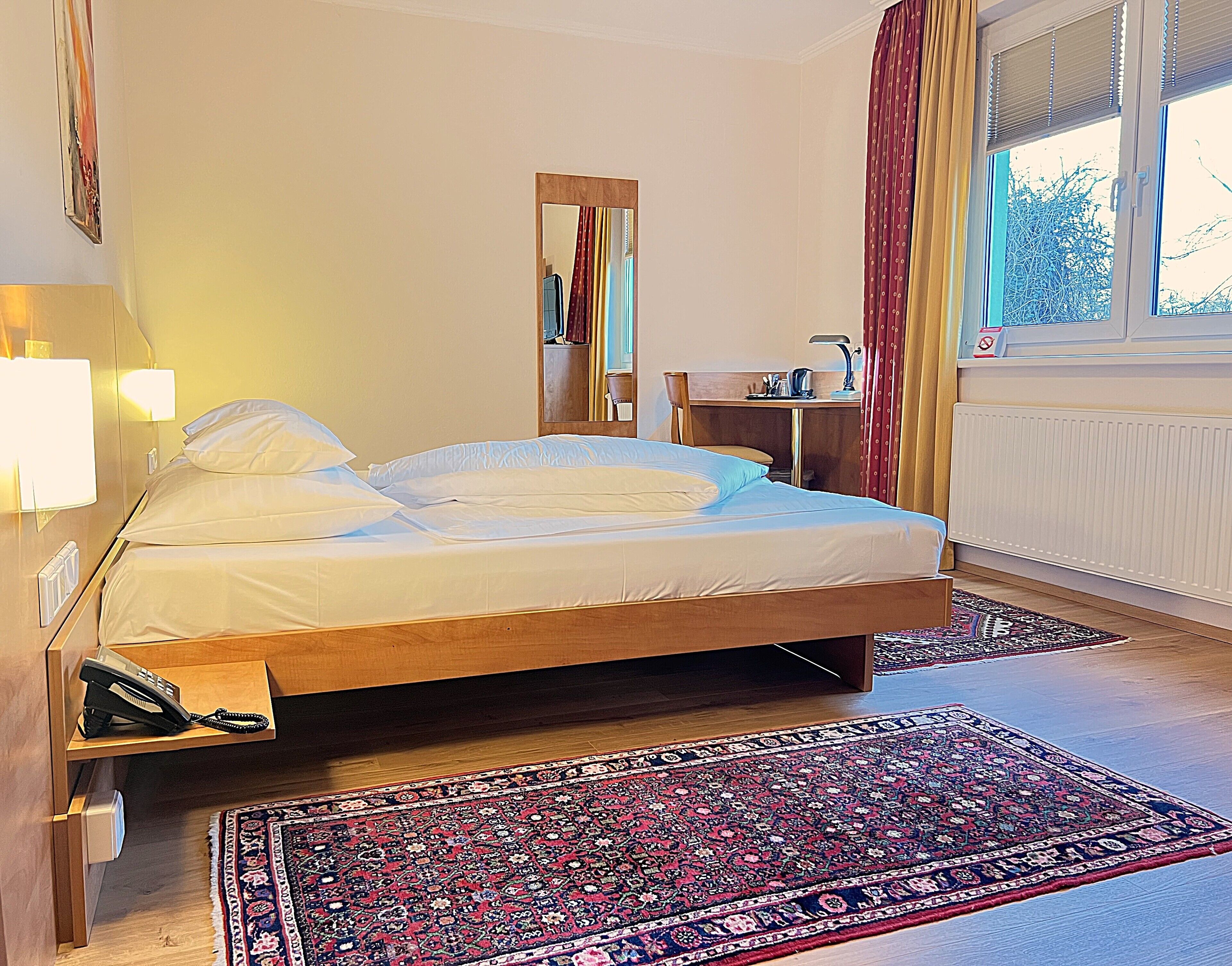 A bedroom with a bed, a desk, a mirror, a radiator, and a window with curtains.