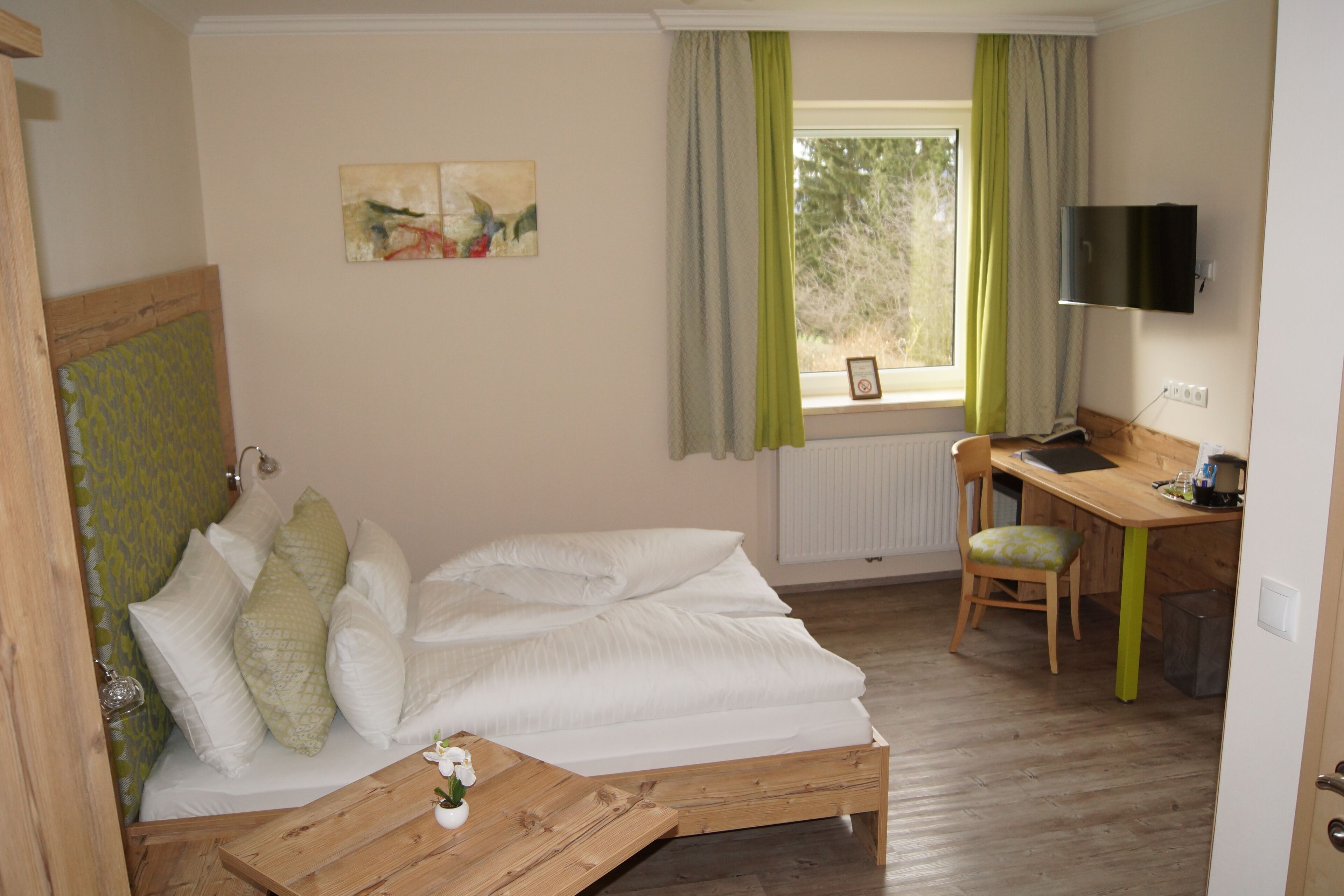 Een slaapkamer met een bed, bureau en stoel. Er is een raam met gordijnen, een schilderij aan de muur en een aan de muur bevestigde televisie.